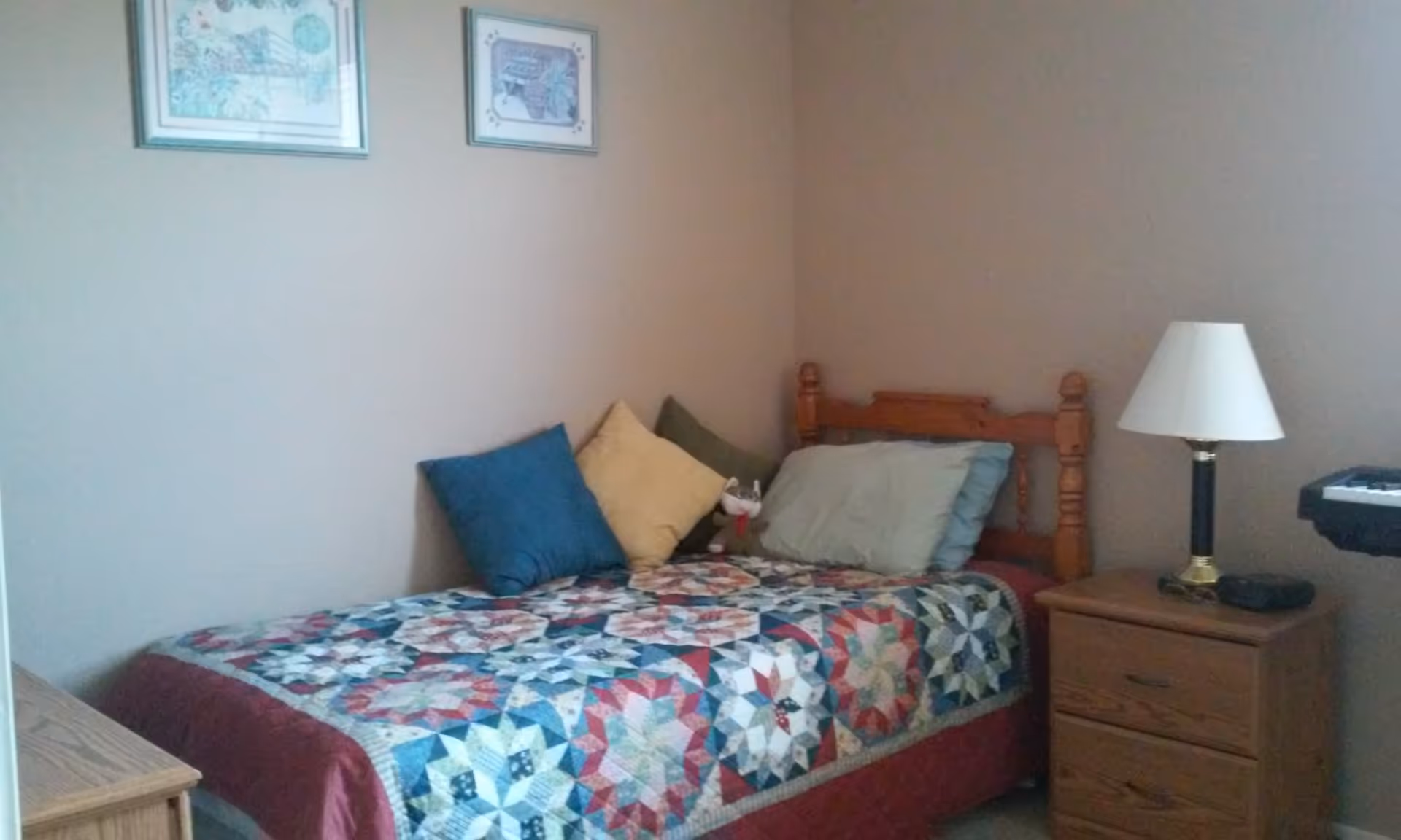 A small bedroom with a single wooden bed covered with a colorful patchwork quilt and several pillows. There is a wooden nightstand next to the bed with a lamp and some items on it. Two framed pictures hang on the beige wall above the bed.