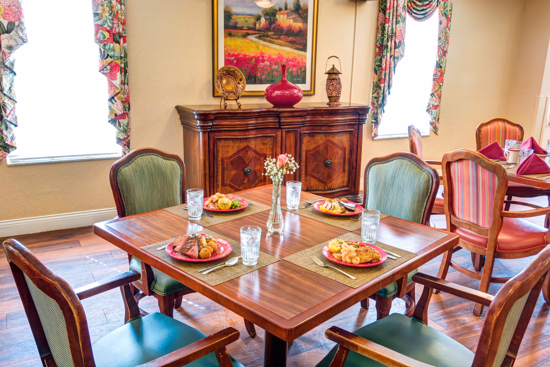 Well-lit dining room with a wooden table set for four with plates of food, glasses, and a decorative cabinet and artwork in the background.