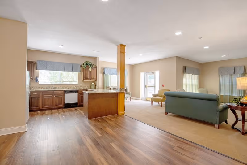 A bright and spacious interior of an assisted living facility showing a kitchen area with wooden cabinets, granite countertops, and a central island. Adjacent to the kitchen is a living room area with a green sofa, an armchair, a side table with a lamp, and large windows with curtains allowing natural light to fill the space.