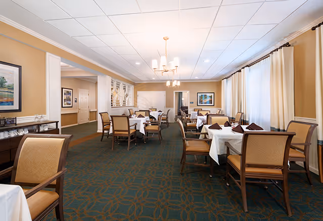 A dining room with several tables covered in white tablecloths, each set with folded brown napkins and chairs around them. The room has beige walls, large windows with cream curtains, framed artwork on the walls, and ceiling lights providing warm illumination.