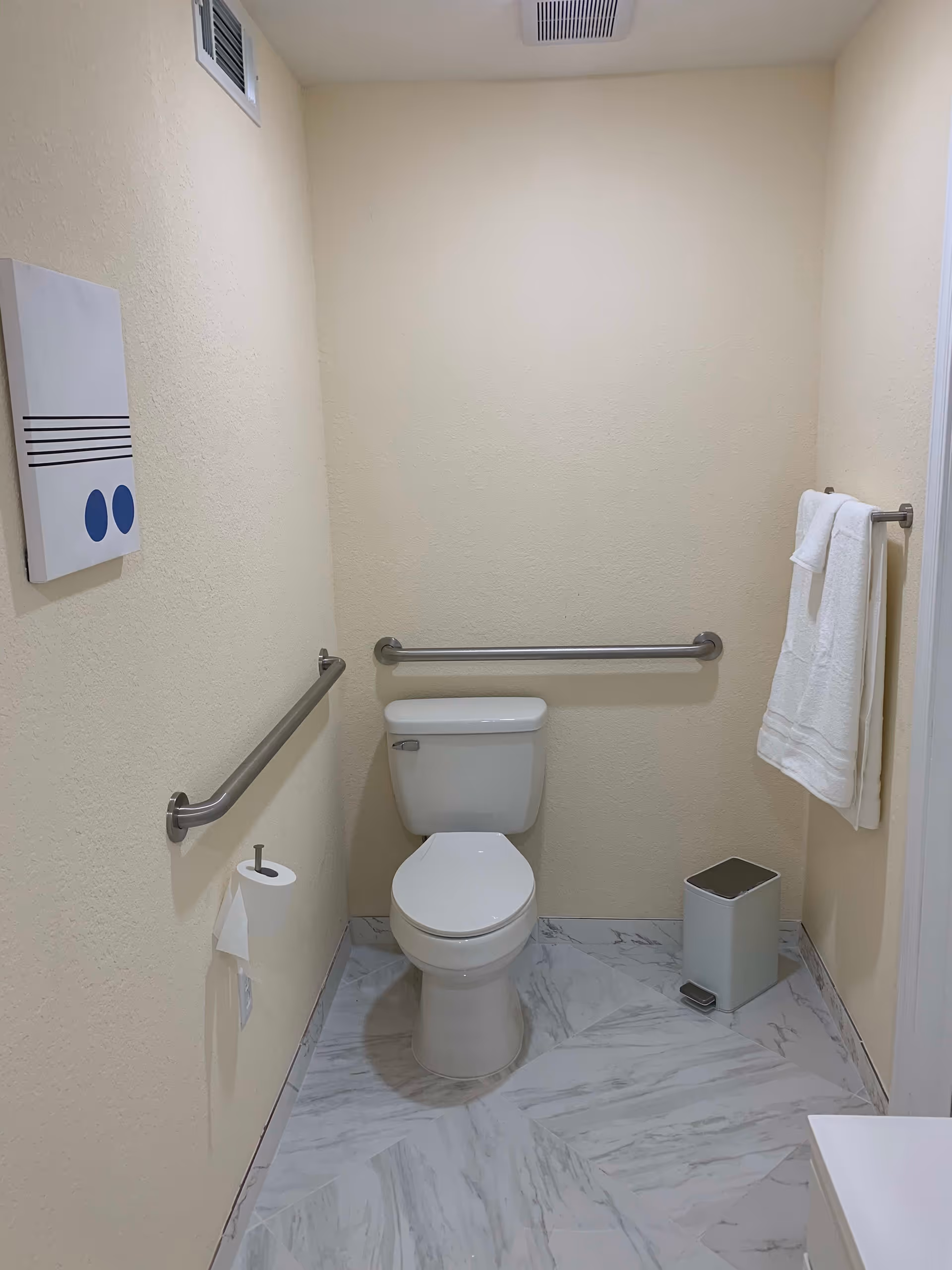 A clean, accessible bathroom with a white toilet centered against a beige wall. There are stainless steel grab bars on the left and back walls, a toilet paper holder with a roll on the left wall, a small white trash can with a foot pedal on the floor to the right, and two white towels hanging on a towel rack on the right wall. The floor is covered with white and gray marble-patterned tiles, and there is a simple piece of wall art with blue circles on the left wall.