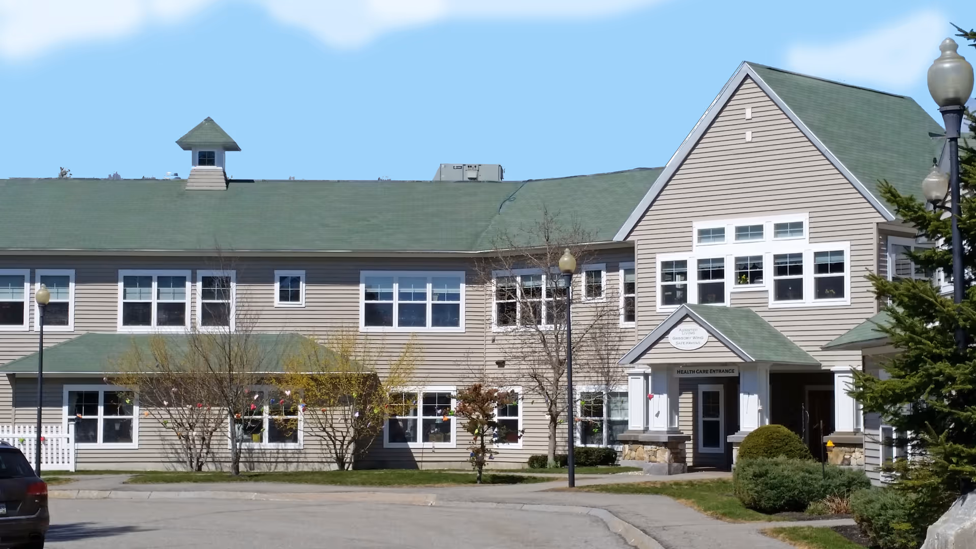 Exterior view of a two-story assisted living facility building with beige siding and a green roof. The building has multiple windows and a main entrance labeled 'Health Care Entrance'. There are trees and shrubs in front of the building, along with street lamps and a paved driveway.