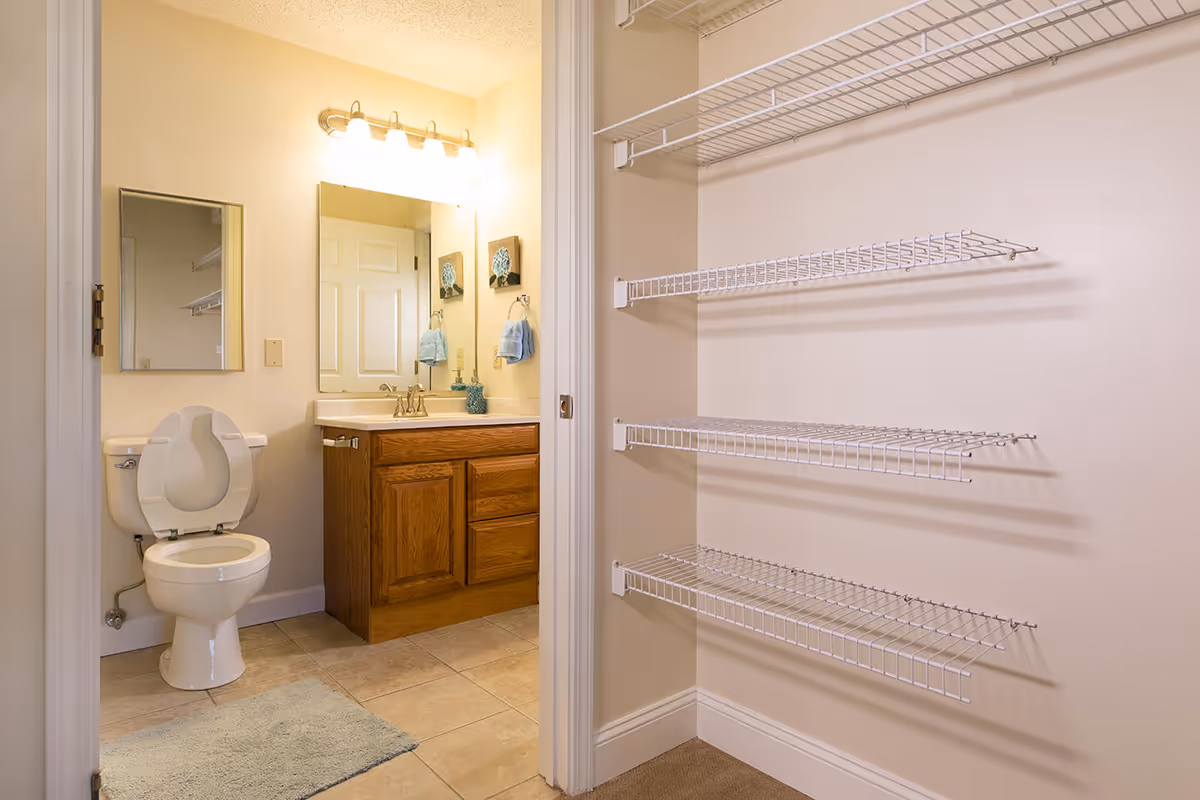 View into a bathroom showing a toilet with raised seat, a wood vanity with mirror and lights, and an adjacent closet with white wire shelving.