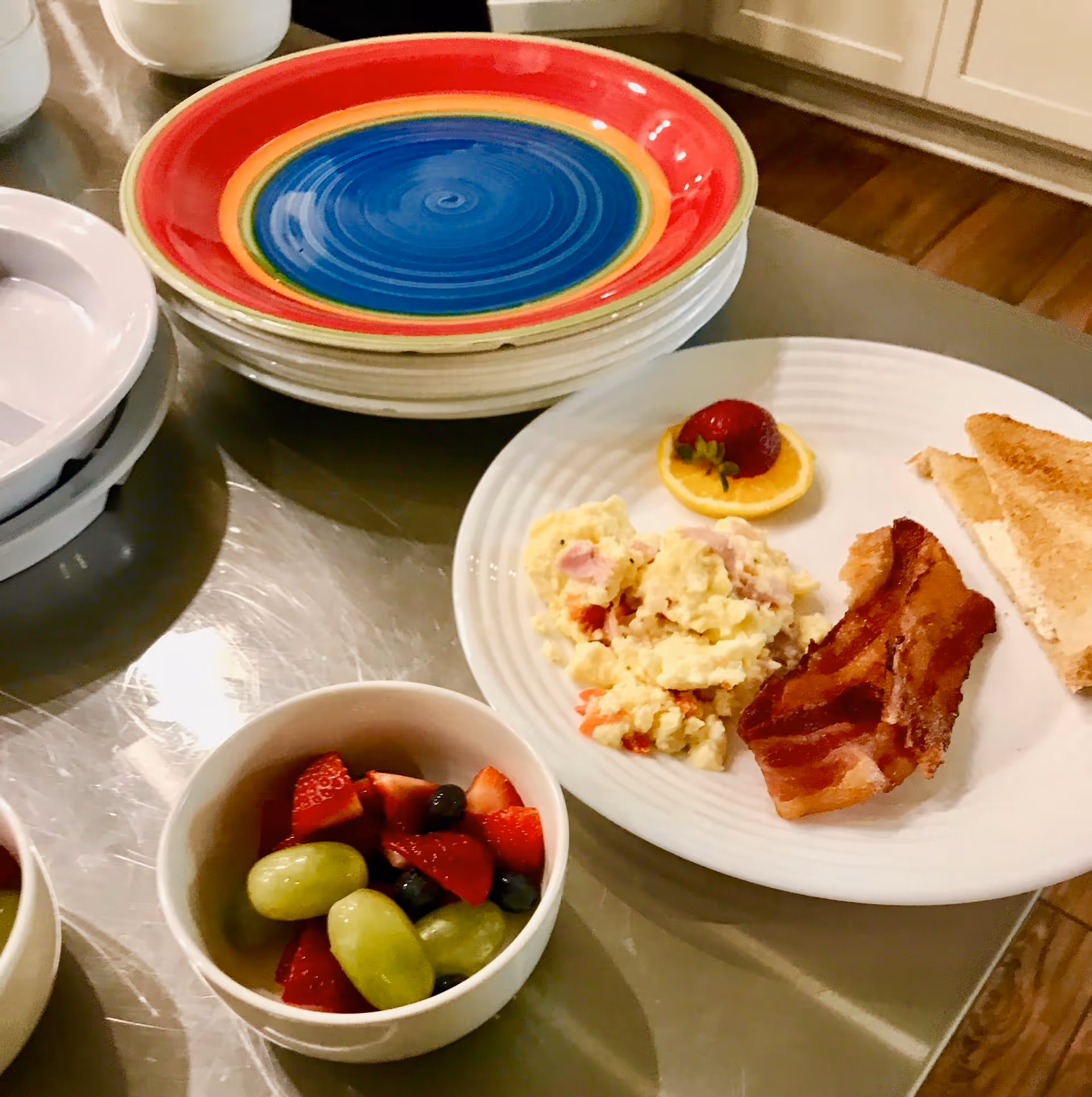 A breakfast plate with scrambled eggs mixed with vegetables, two strips of bacon, a slice of toast with butter, and a garnish of a strawberry on an orange slice. Next to the plate is a bowl of mixed fruit including green grapes, strawberries, and blueberries. In the background, there is a stack of colorful plates on a kitchen counter.