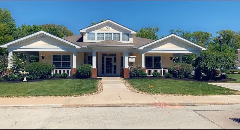 Front exterior view of a single-story senior living facility building with a covered entrance, white columns, and a well-maintained lawn under a clear blue sky.