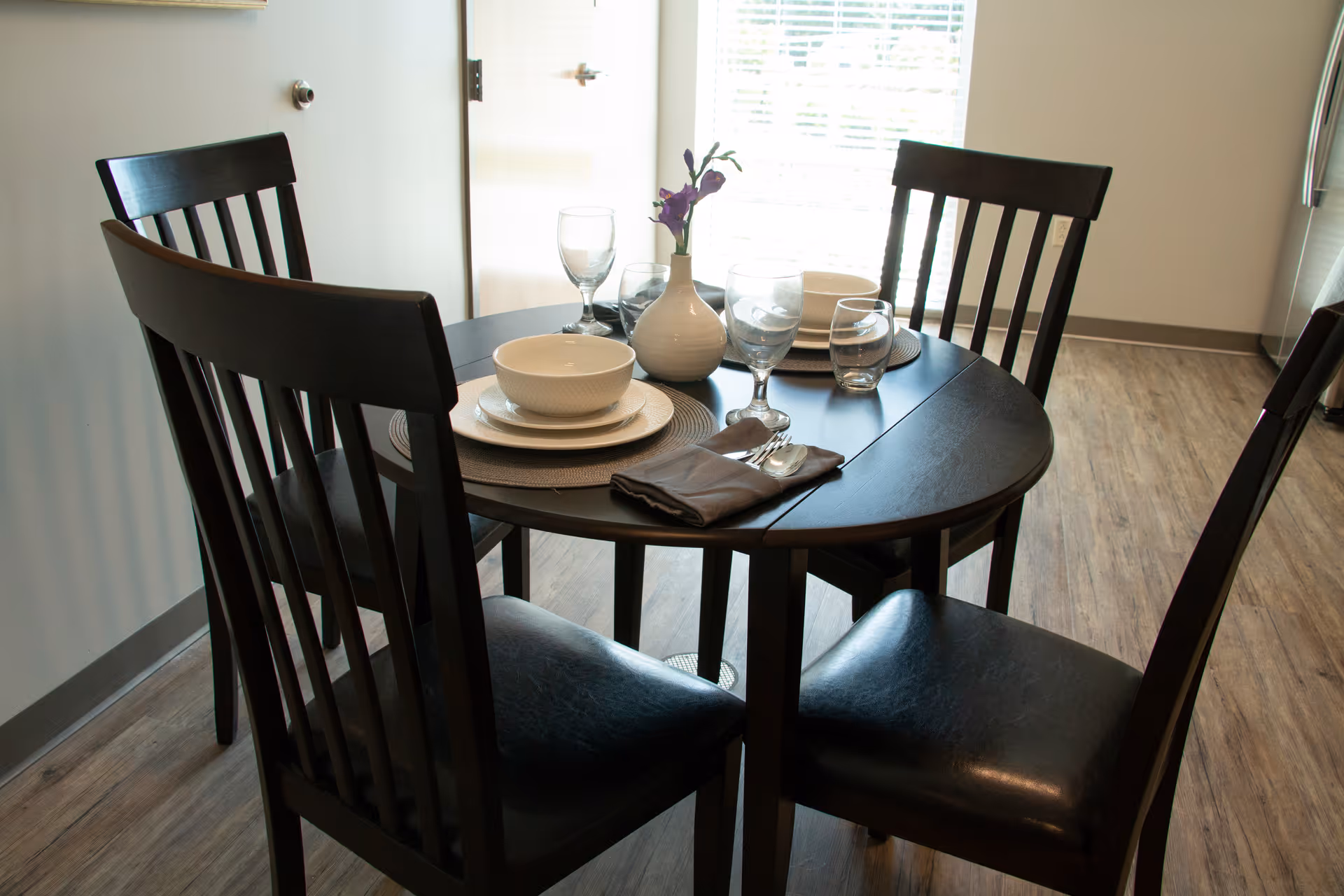 A dark wooden round dining table set for four with matching chairs. The table is set with white bowls, plates, glasses, silverware wrapped in a napkin, and a small white vase with purple flowers. The room has light-colored walls and wood flooring, with a window covered by blinds in the background.