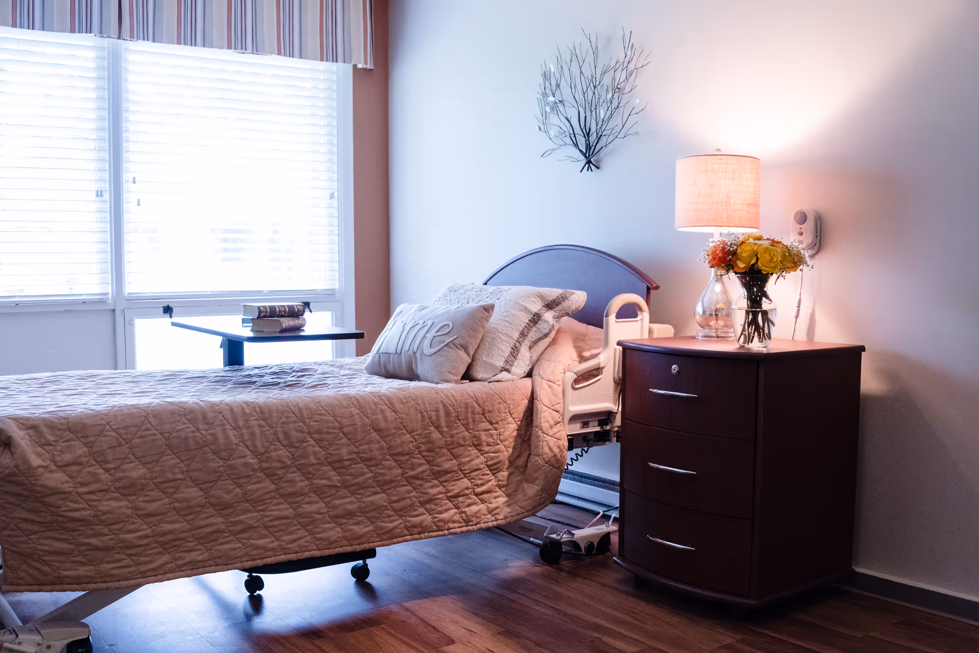 A furnished bedroom with a hospital-style bed, bedside table holding a lamp and vase of flowers, and a large window with blinds.
