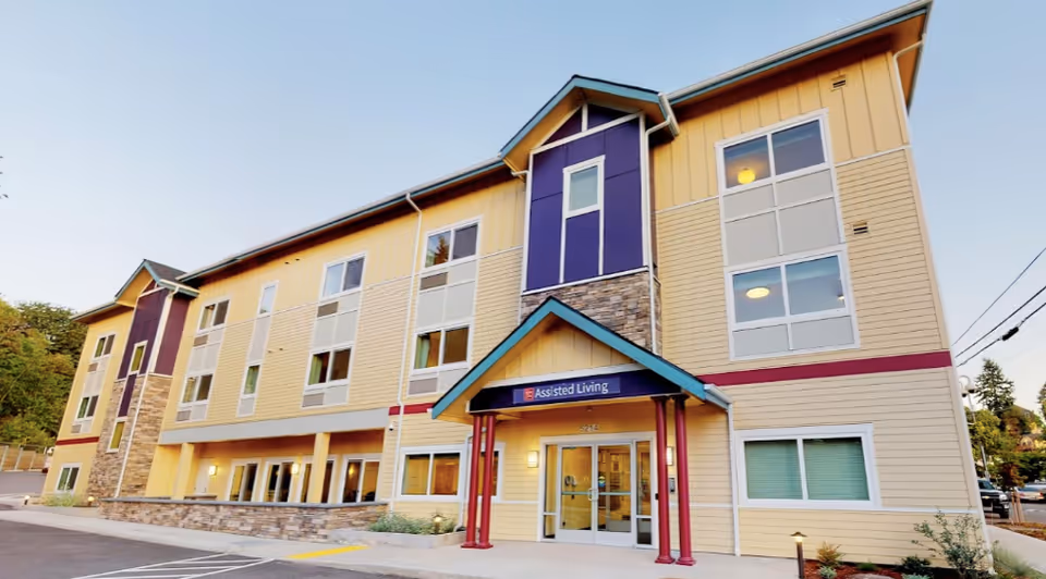 Exterior view of a three-story assisted living facility building with beige and purple siding, stone accents, and a covered entrance with a sign that reads 'Assisted Living'. The building has multiple windows and is surrounded by a paved parking area and some greenery.