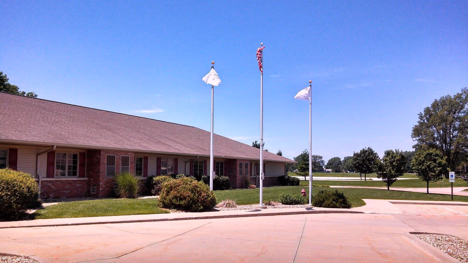 Exterior view of a single-story brick and siding building with a sloped roof, surrounded by green grass, bushes, and trees under a clear blue sky. Three flagpoles with flags, including an American flag, stand in front of the building near a paved driveway and parking area.