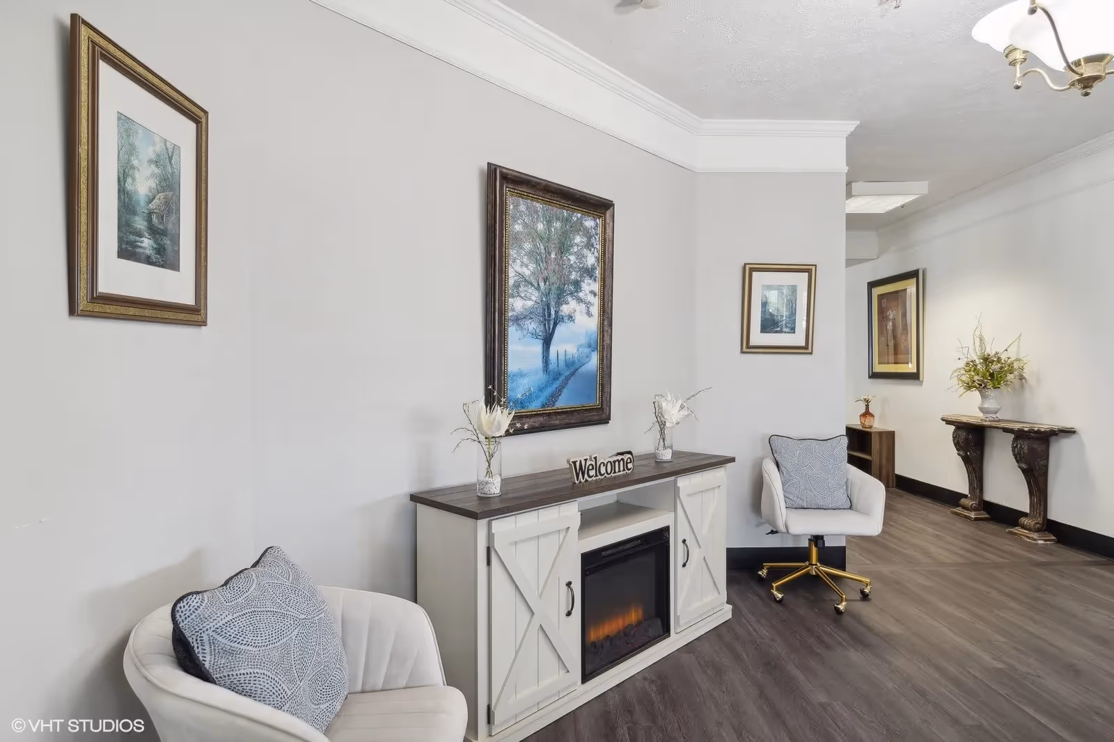 A cozy interior seating area with two white cushioned chairs, each with a decorative pillow. Between the chairs is a white cabinet with a dark wooden top, featuring a built-in electric fireplace and a 'Welcome' sign on top. Above the cabinet hangs a framed painting of a tree-lined path. The walls are light-colored and adorned with additional framed artwork. The floor is dark wood, and there is a decorative console table with a vase of flowers in the background.