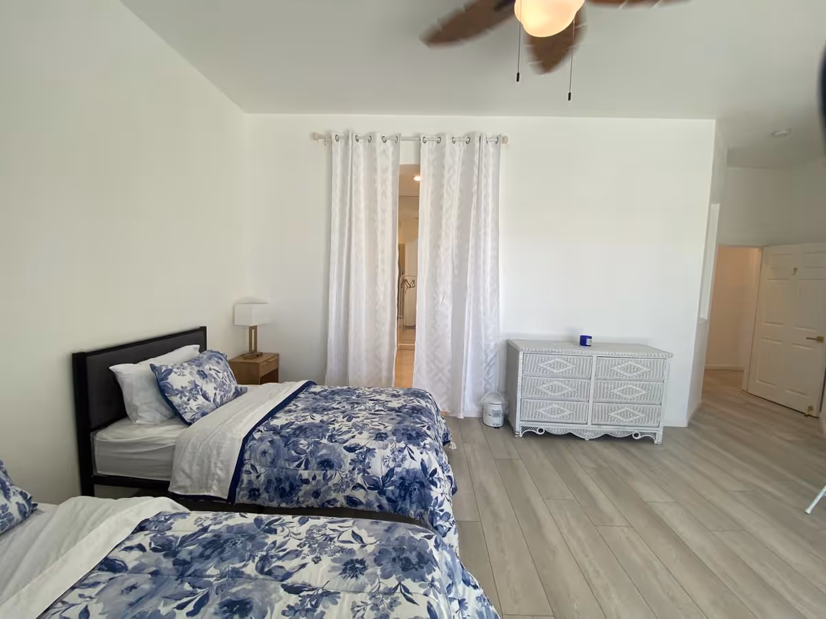A bright bedroom with two twin beds featuring blue and white floral bedding. There is a wooden nightstand with a lamp between the beds. White curtains hang in the center of the far wall, partially covering an open doorway. A white wicker dresser is positioned against the right wall, and the floor is light-colored wood. A ceiling fan with a light is visible at the top of the image.