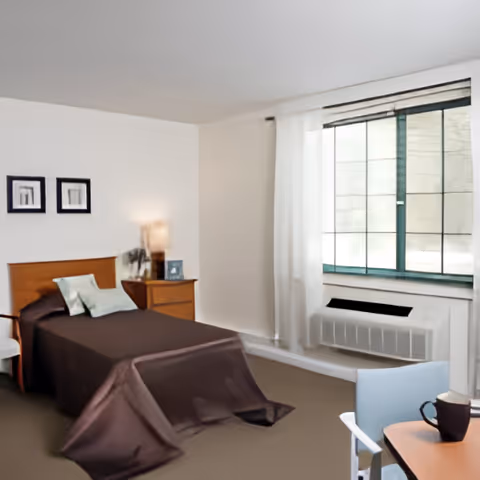 A simple bedroom with a single bed covered in a dark brown blanket, two pillows, a wooden headboard, and a wooden nightstand with a lamp and a small framed picture. There are two framed pictures on the wall above the bed. A window with white curtains and a heating/cooling unit below it is on the right side. A small table with a chair and a cup is partially visible in the foreground.