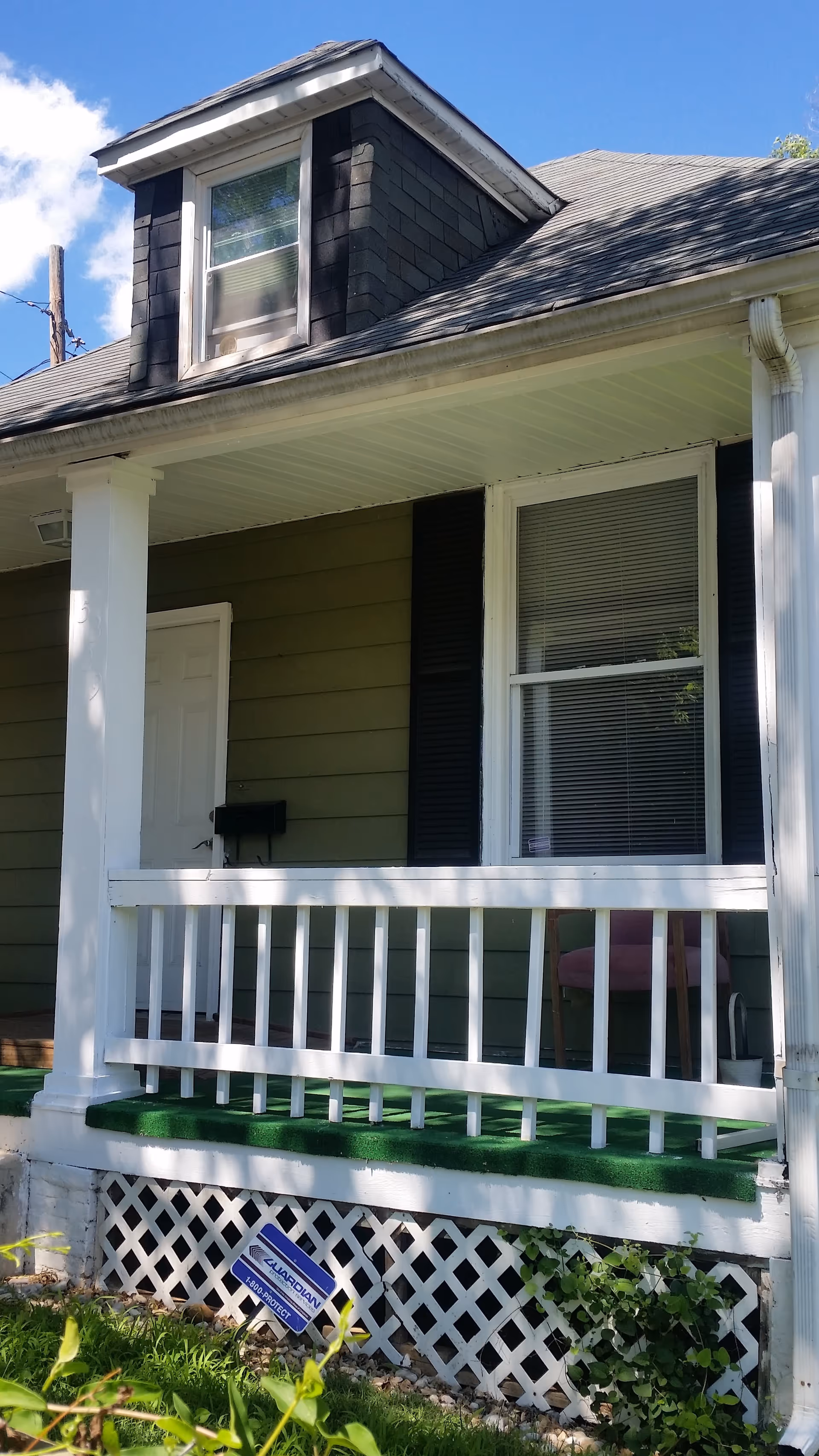 Front porch of a small house with white railing, green siding, a window with blinds, and a dormer above.
