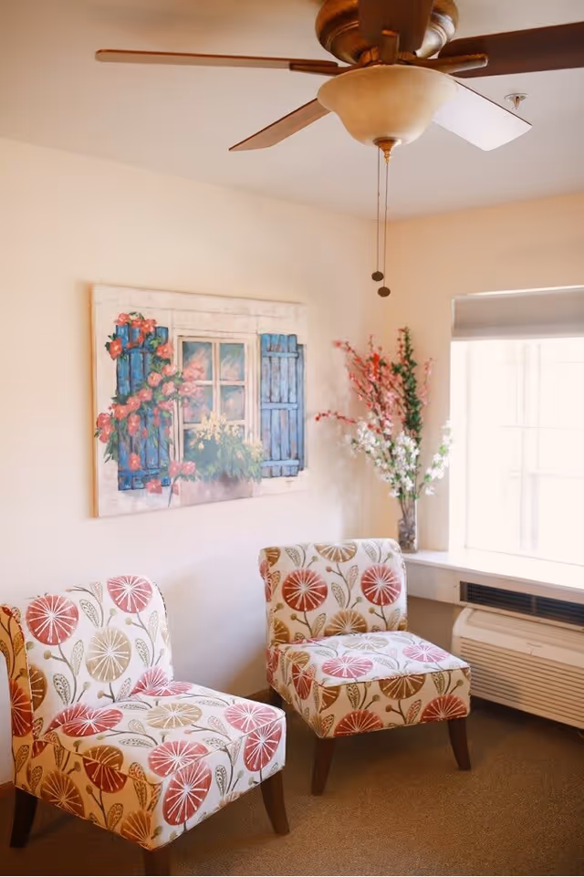 Two patterned accent chairs beside a window with floral artwork, a vase of flowers, and a ceiling fan overhead.