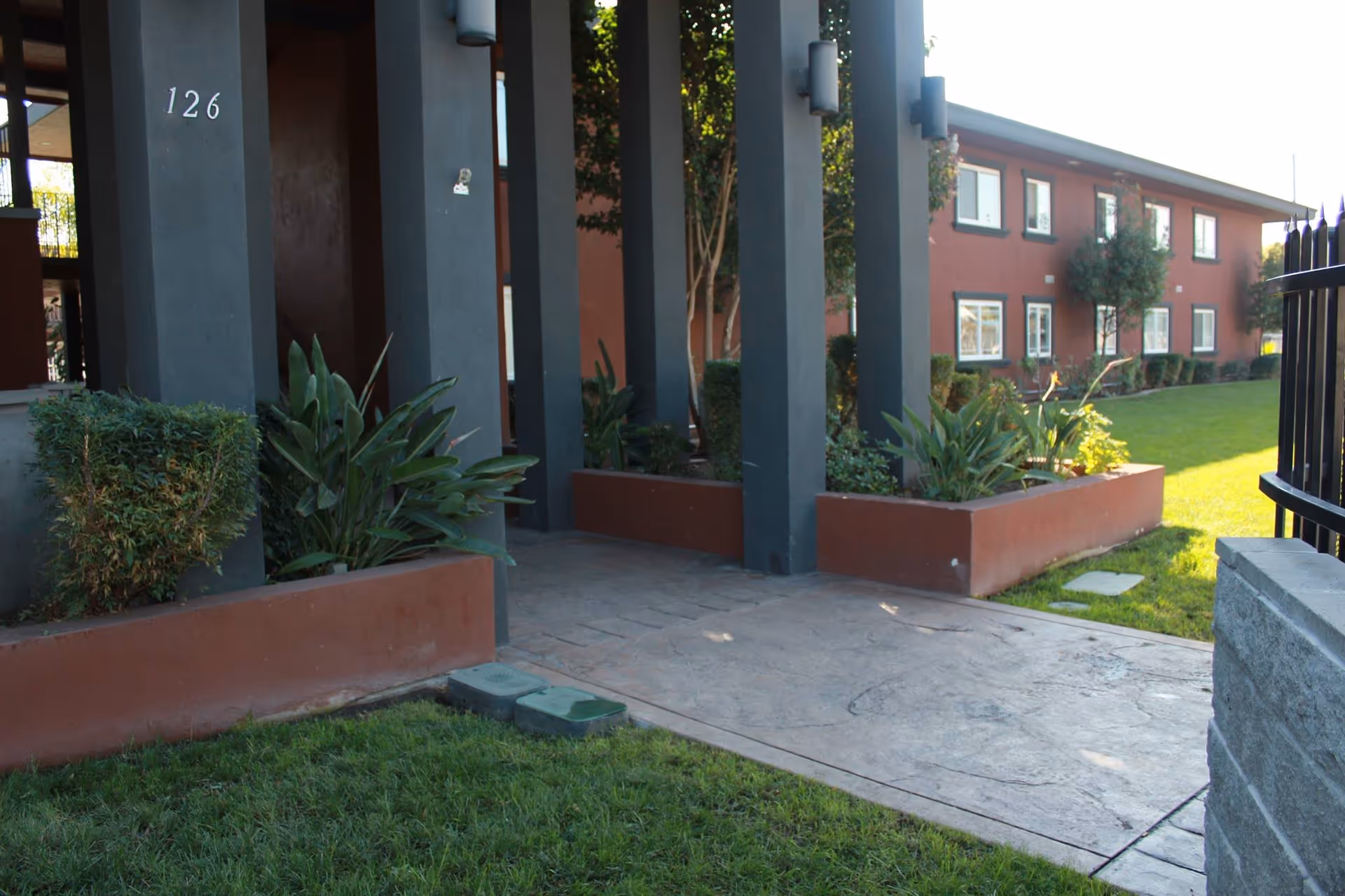 Outdoor view of the entrance area of a building with the number 126 on a dark pillar. The scene includes a paved walkway, green grass, plants in raised planters, and a two-story red building with multiple windows in the background.
