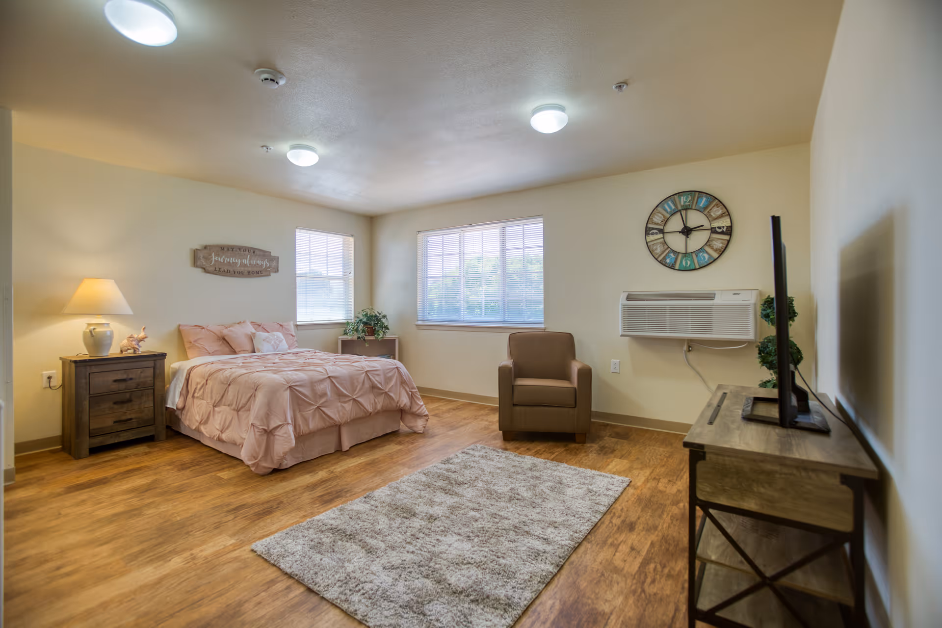 A cozy bedroom with a bed covered in a pink comforter and pillows, a wooden nightstand with a lamp and decorative items, a brown armchair, a wall-mounted air conditioner, a colorful wall clock, two windows with blinds, a wooden floor, and a beige rug in the center.