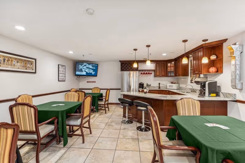 Interior view of a common area with a kitchen and dining space. The room features several tables covered with green tablecloths and surrounded by striped cushioned chairs. The kitchen area has wooden cabinets, a marble countertop with bar stools, a refrigerator, microwave, coffee machine, and pendant lights. A TV is mounted on the wall, and there is a decorative sign that reads 'COCKTAILS' on the right side.