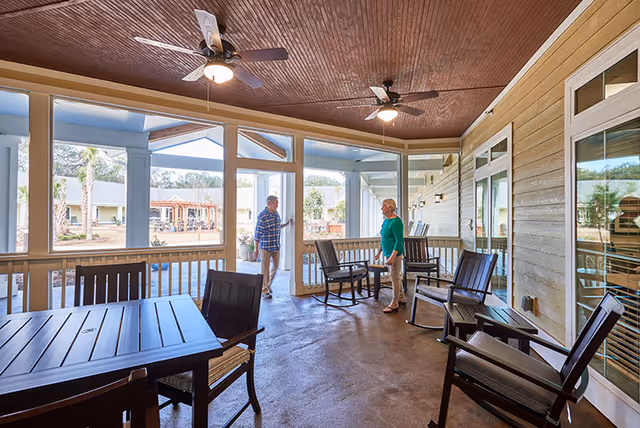 Covered outdoor patio area with ceiling fans, several chairs, and tables. Two people are standing and talking near the entrance to the patio. The background shows a garden and other buildings.
