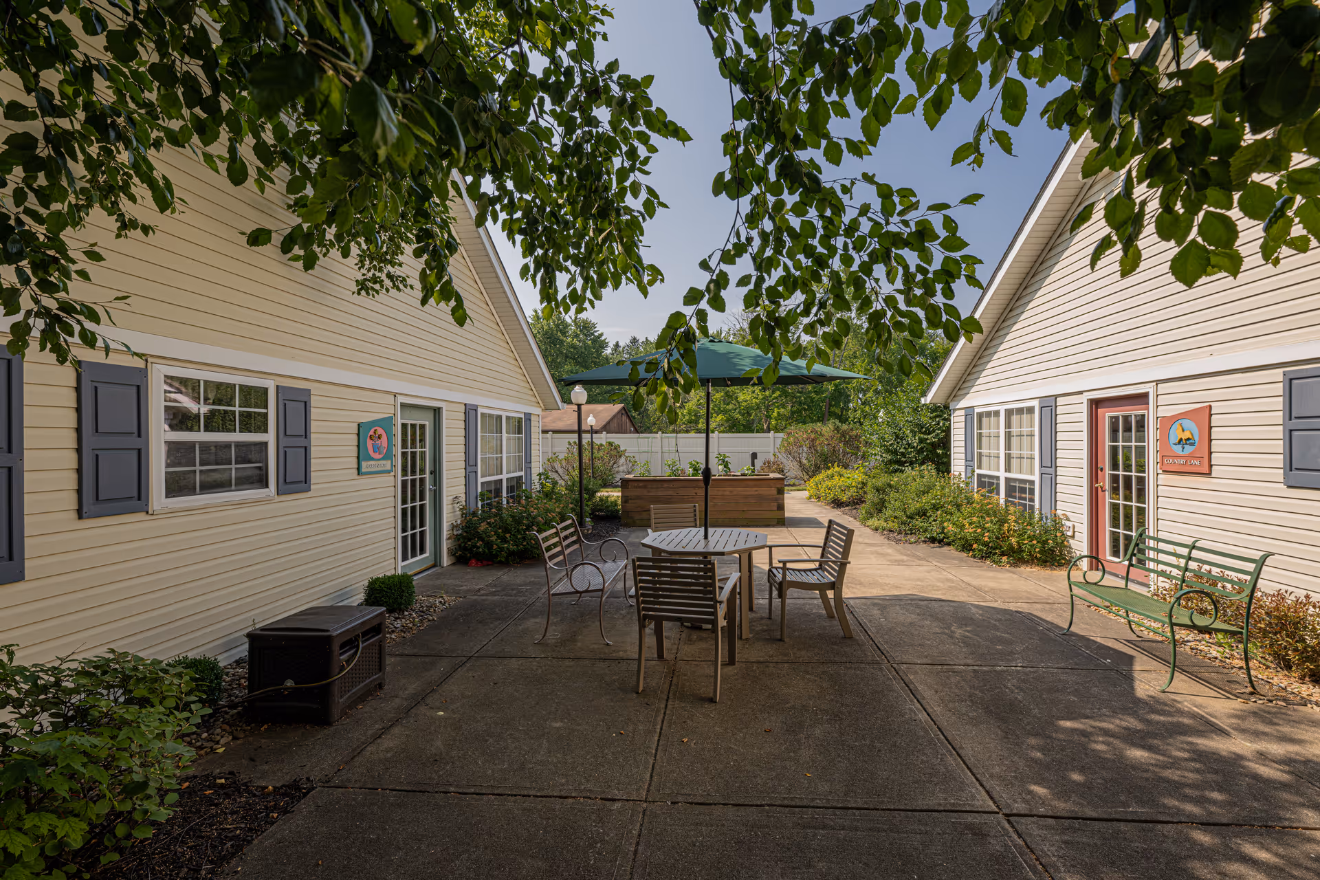 A shaded courtyard between two single-story buildings with a table, chairs, umbrella and benches on a paved patio.