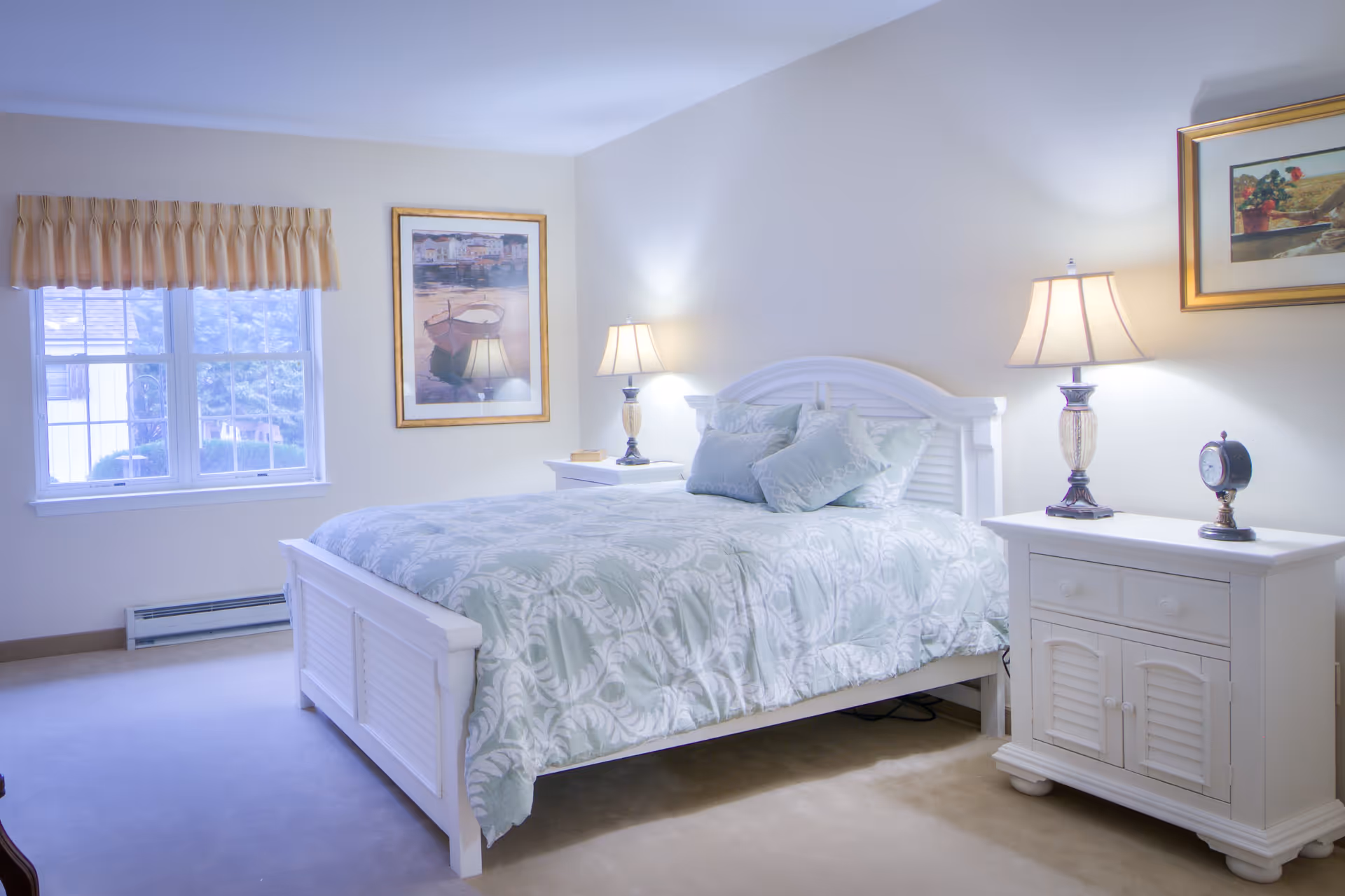 A bright bedroom with a white wooden bed frame and matching nightstands on either side. The bed is made with a light blue and white patterned comforter and several pillows. Each nightstand has a lamp with a beige shade, and one has a small clock. There are two framed pictures on the walls and a window with a beige valance letting in natural light.