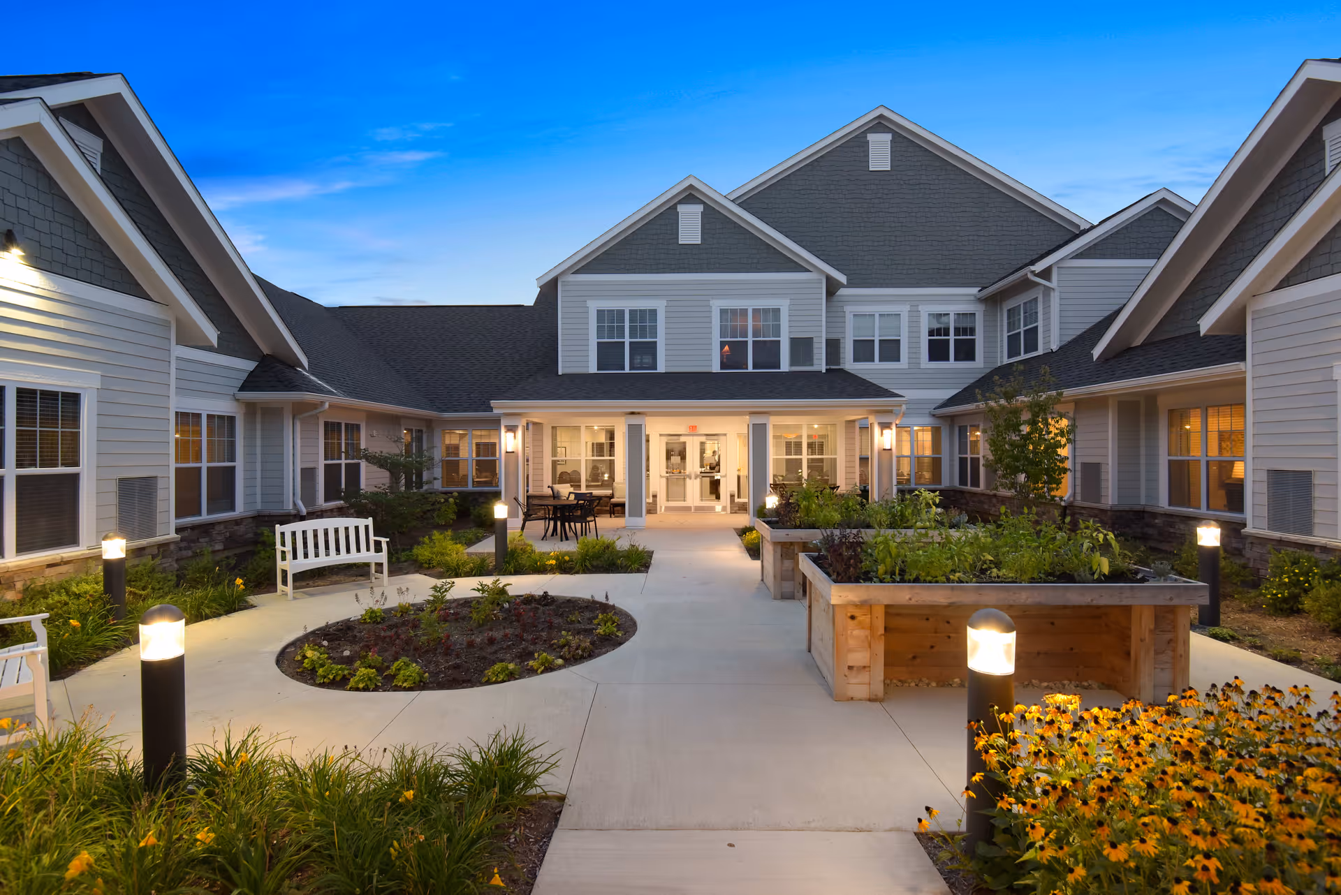 Exterior view of American House Hampton Village at dusk, showing a well-lit courtyard with benches, raised garden beds, and a pathway leading to the entrance of a two-story building with multiple windows.