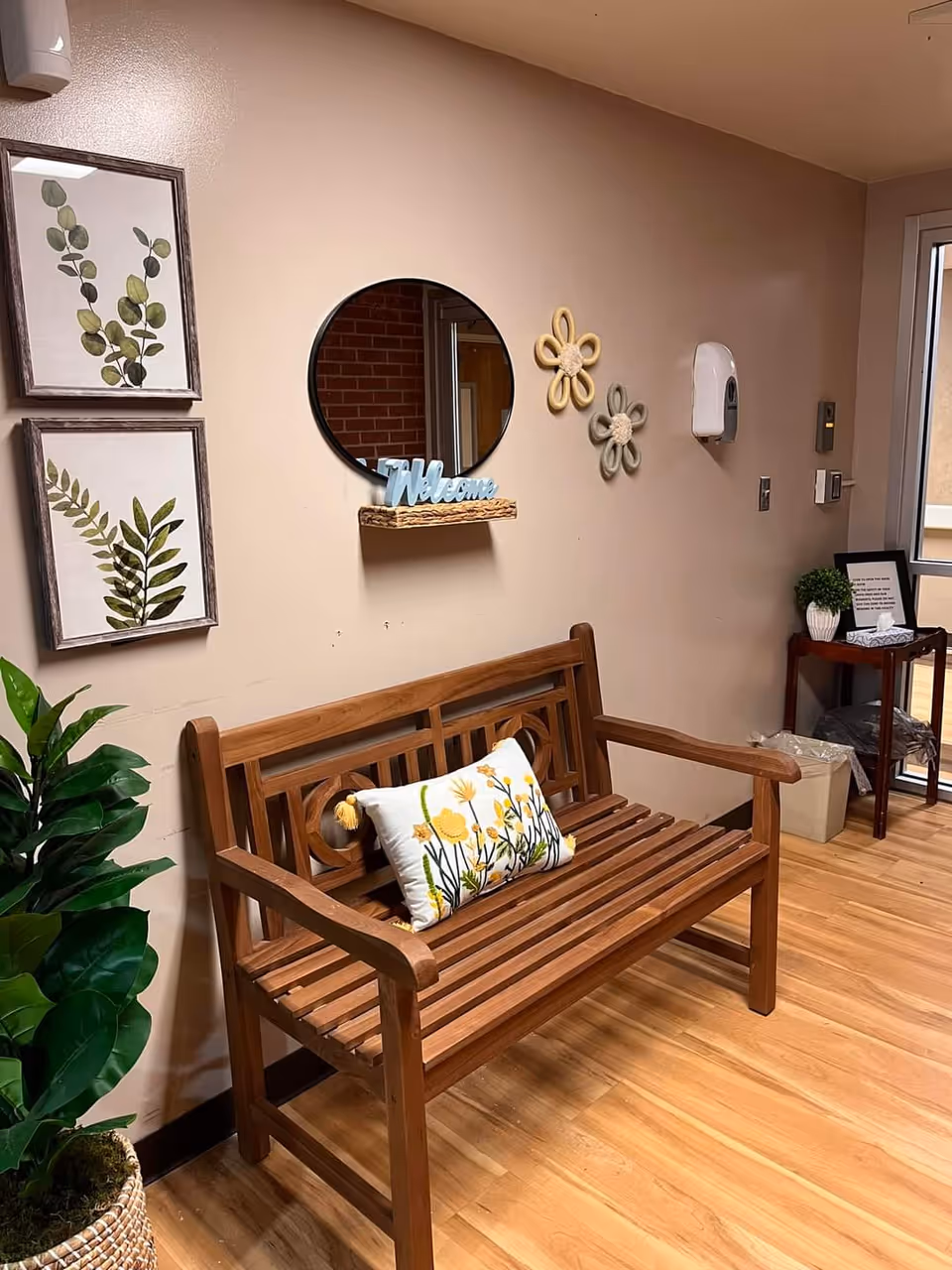 A cozy indoor seating area with a wooden bench that has a decorative pillow featuring yellow flowers. Above the bench, there is a round mirror with a small shelf holding a blue 'Welcome' sign. The wall is decorated with two framed botanical prints and two flower-shaped wall hangings. To the left of the bench is a potted green plant, and to the right is a small wooden table with a framed sign, a small plant, and a tissue box. The floor is wooden, and there is a hand sanitizer dispenser mounted on the wall.