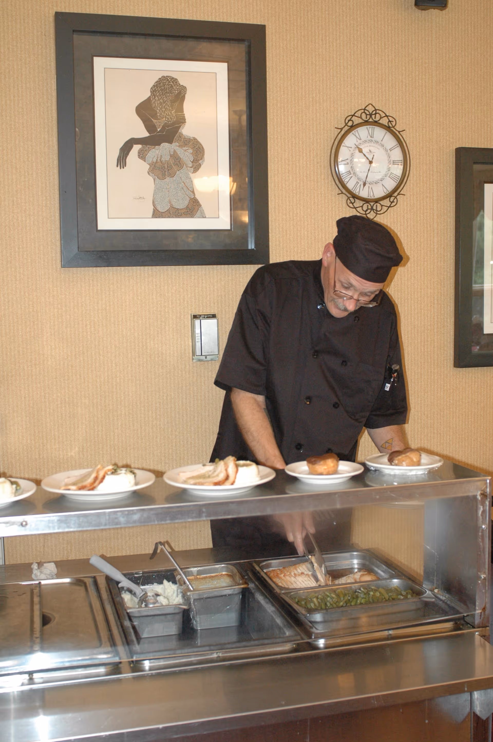 A chef dressed in black uniform and hat is serving food behind a cafeteria-style serving counter. There are plates with food items such as mashed potatoes, green beans, and bread rolls on the counter. The background features a beige wall with a framed artwork and a decorative clock.