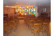Interior view of a room with rows of wooden chairs facing a stage area. The back wall features a colorful stained glass window. The room is well-lit with ceiling lights and has framed pictures on the walls.