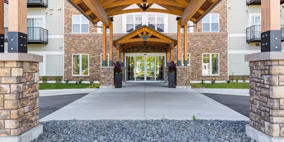 Covered main entrance to a senior living building with wooden beams, stone pillars, a concrete drive, and glass double doors.