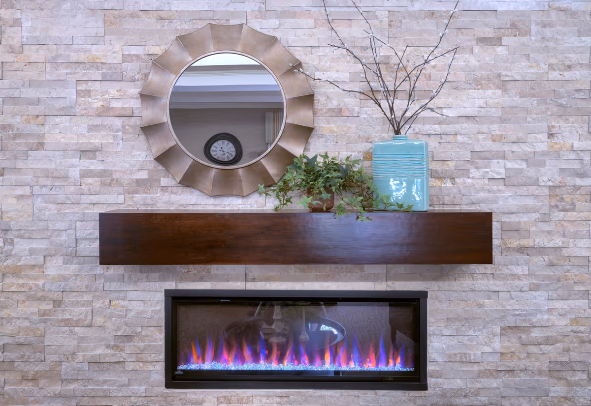 A modern electric fireplace with blue and purple flames set into a stone wall. Above the fireplace is a dark wooden mantel holding a green leafy plant and a large turquoise vase with bare branches. A round decorative mirror with a sunburst frame hangs on the wall above the mantel, reflecting a clock and part of the ceiling.