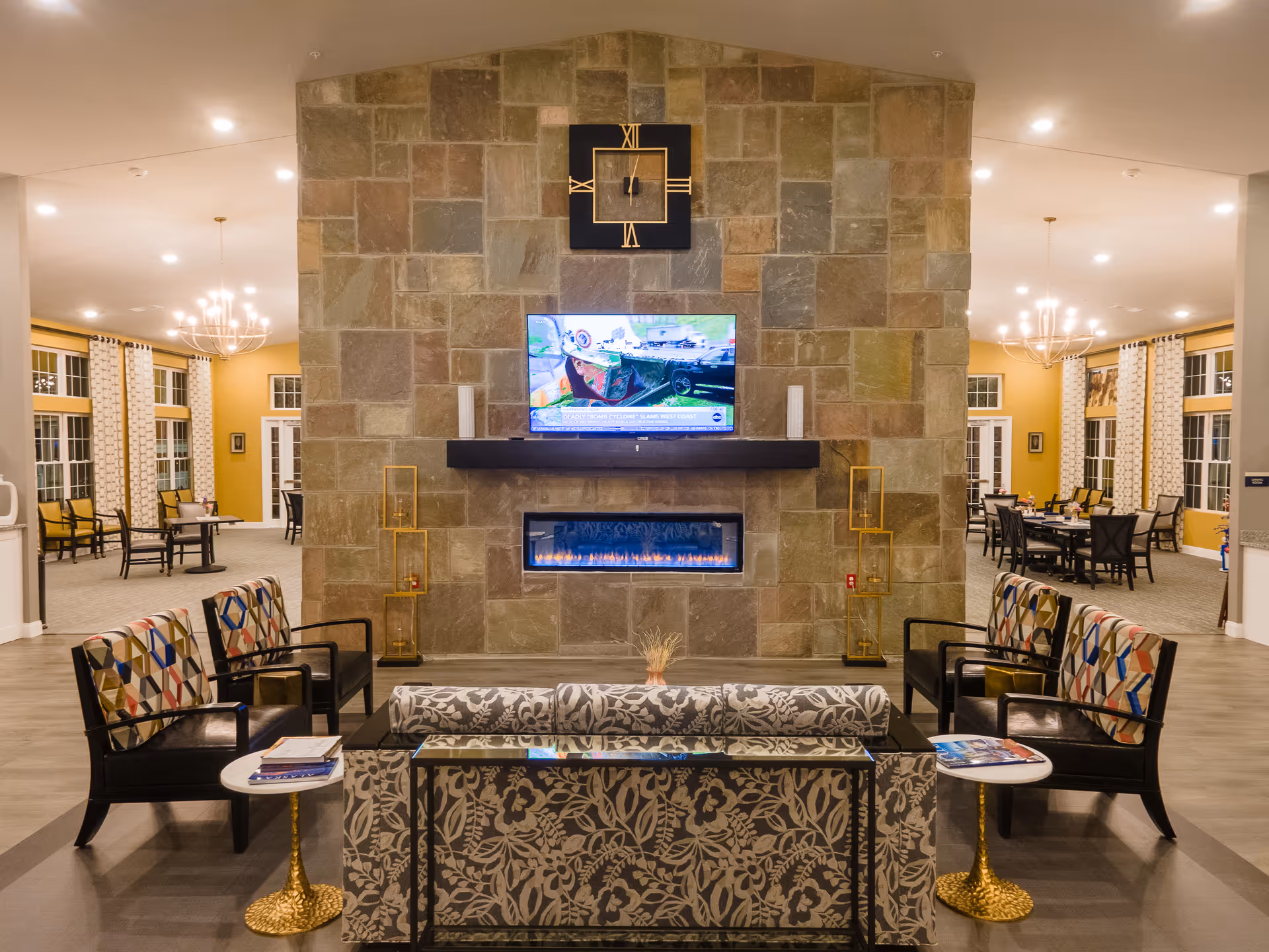 Spacious assisted living lounge with a stone fireplace and wall-mounted TV, seating grouped around a coffee table and dining tables visible in the background.