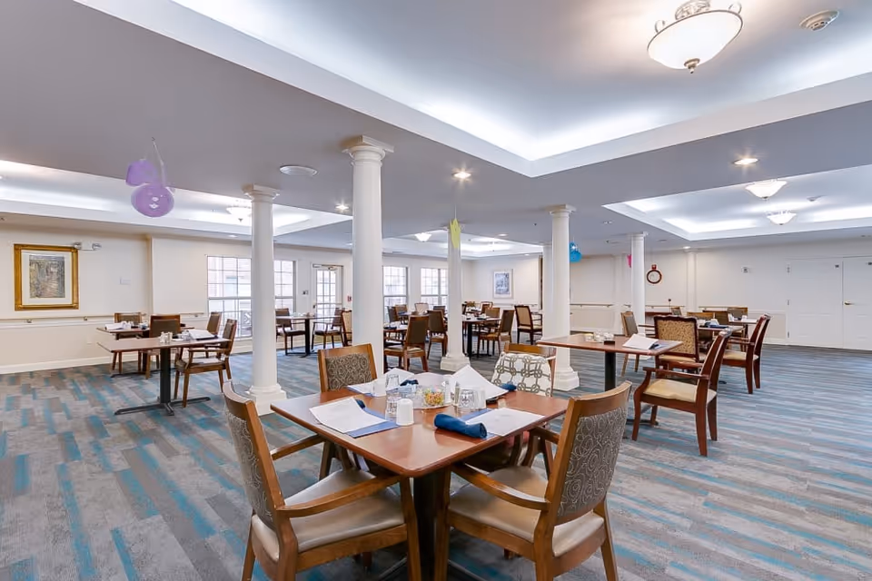 A spacious dining room in a senior living facility with multiple wooden tables and chairs arranged neatly. The room features white columns, large windows letting in natural light, and ceiling lights. Tables are set with napkins, menus, and condiments. The carpet has a blue and gray pattern, and there are framed pictures on the walls.