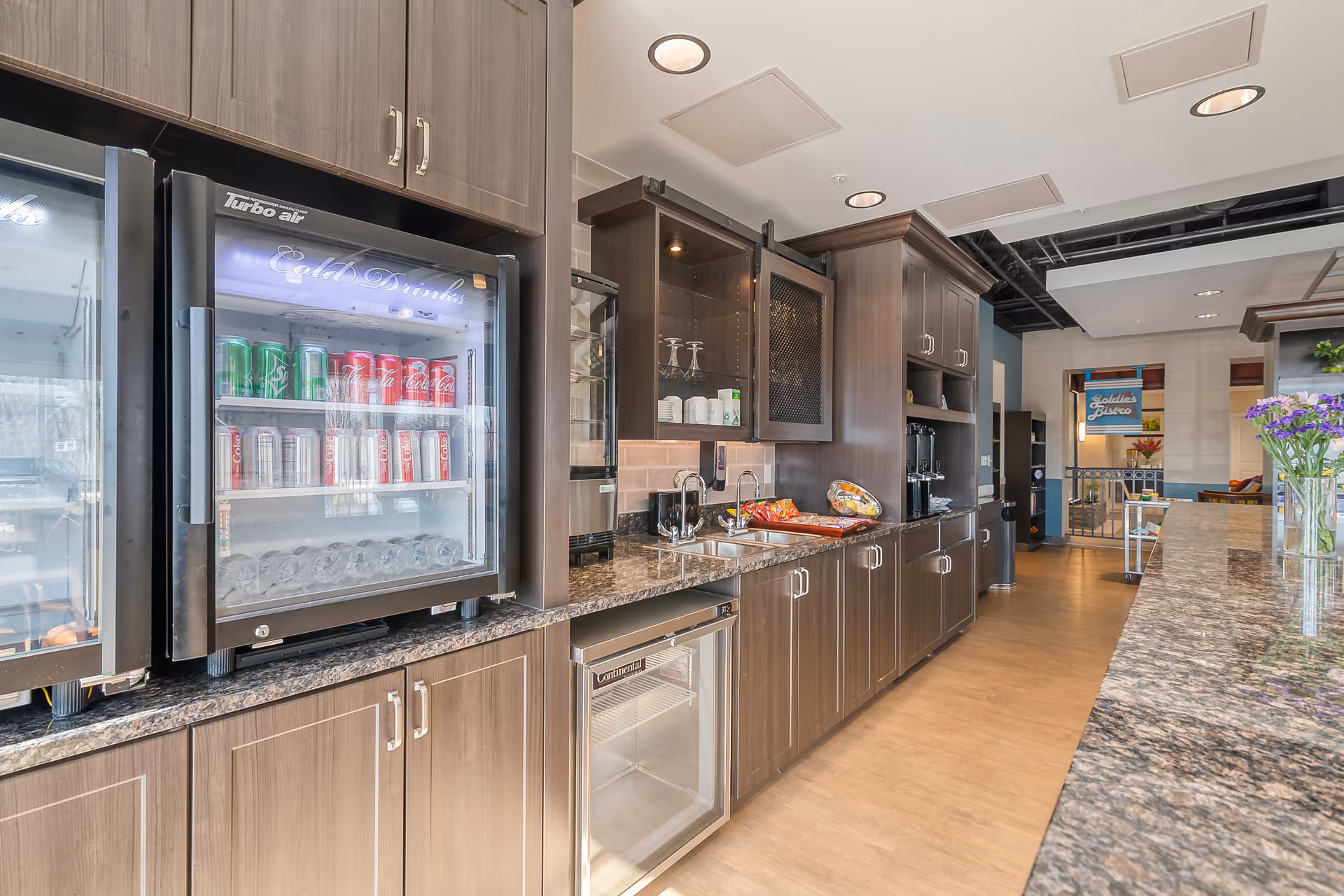 A modern kitchenette/snack bar with glass-front beverage coolers, dark wood cabinets, granite counters, a sink and snacks in a senior living common area.