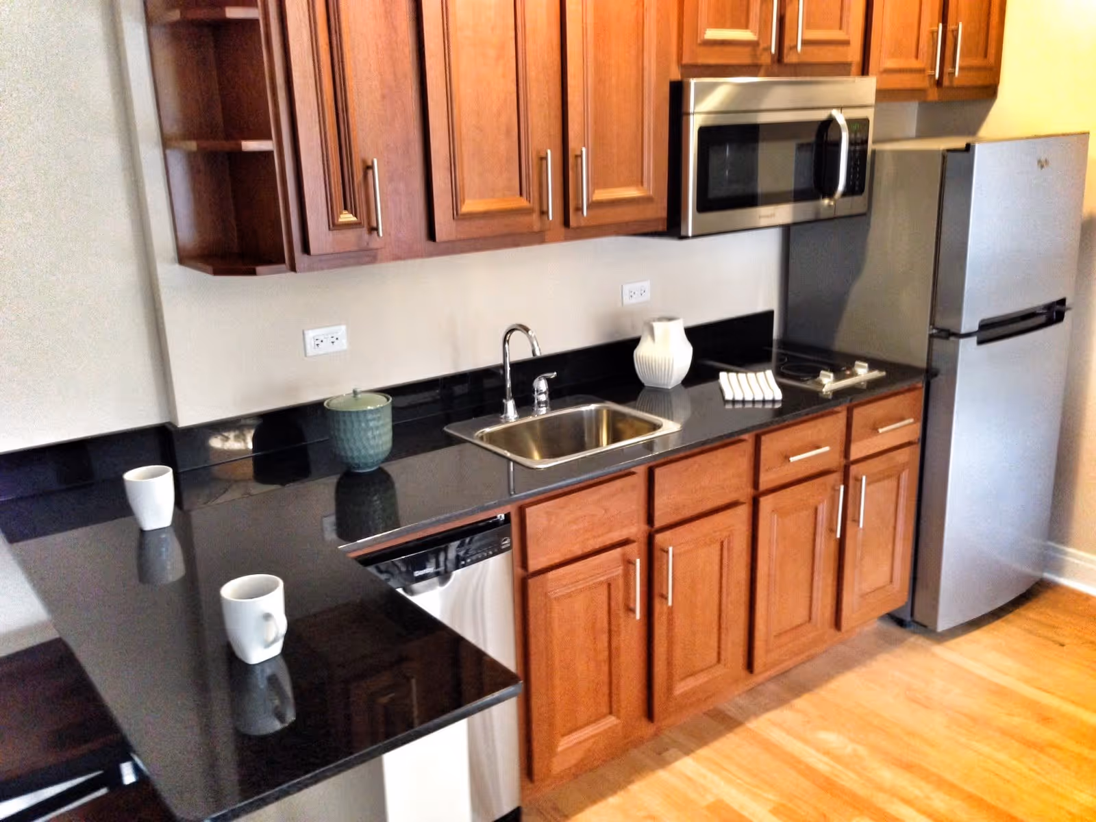 A modern kitchen with wooden cabinets, black granite countertops, a stainless steel sink, a built-in microwave, a refrigerator, and a dishwasher. There are two white mugs, a green container, a white pitcher, and a small stack of white napkins on the countertop. The floor is light wood.