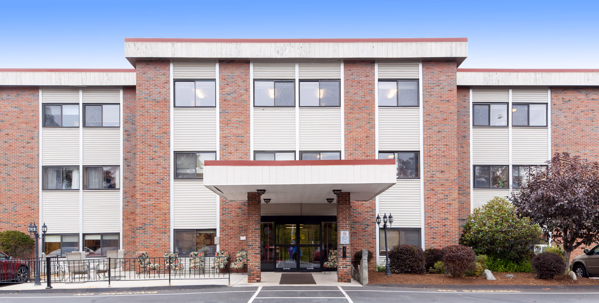 Front exterior of a three-story brick senior living facility with a covered main entrance, windows, and landscaped shrubs.