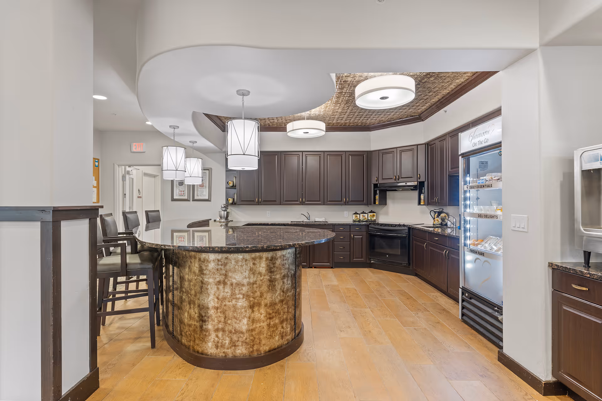 A modern kitchen area in a senior living facility with dark wood cabinets, a curved island with a granite countertop, three pendant lights hanging above the island, and a refrigerator with glass doors labeled 'Glenmoore On the Go'. There are also bar stools around the island and a water dispenser on the right side.