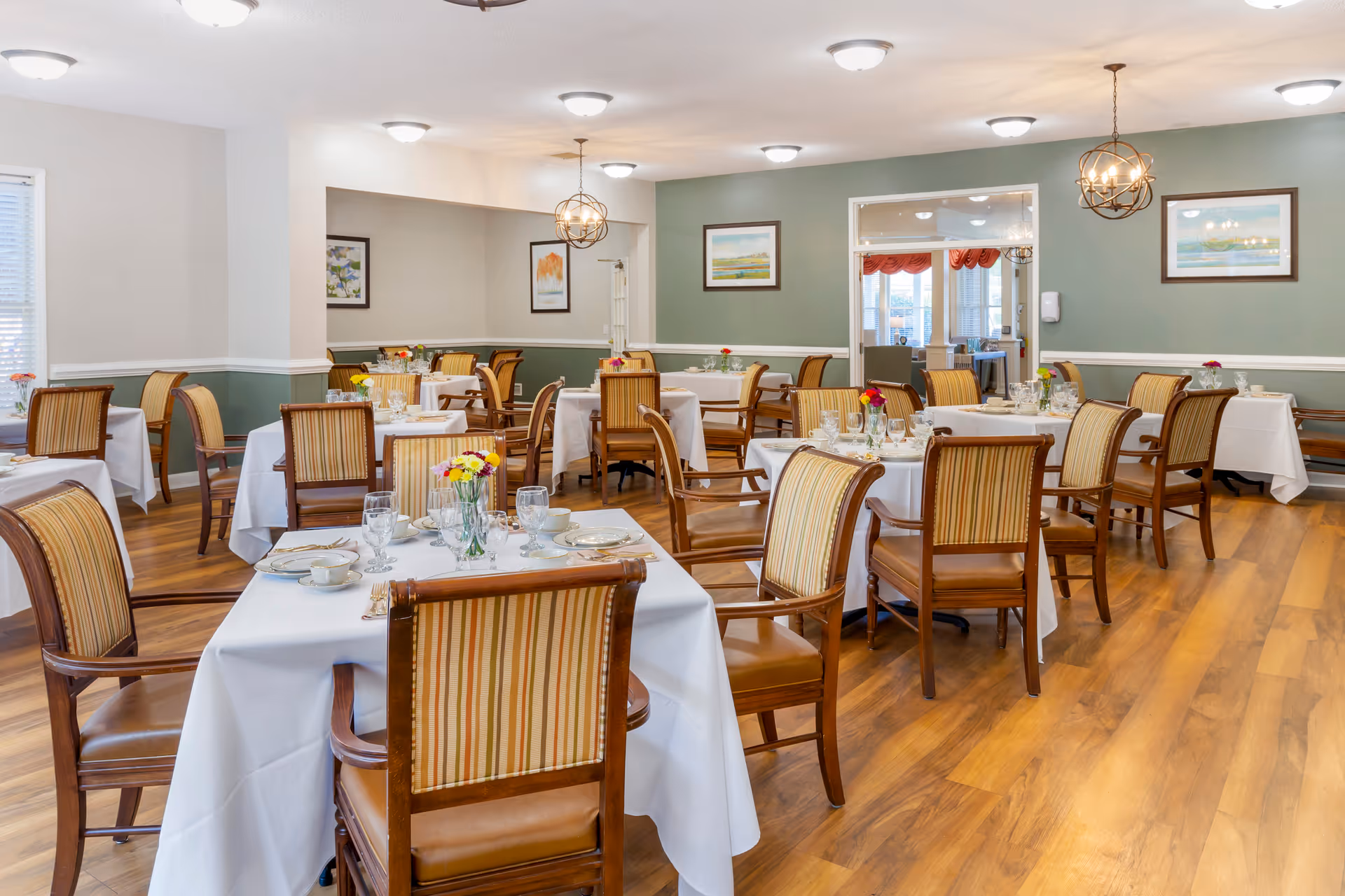 A well-lit dining room with multiple tables covered in white tablecloths, each set with plates, glasses, and silverware. The chairs have wooden frames with striped upholstery. The walls are painted in a combination of light gray and green, adorned with framed artwork. Two spherical pendant lights hang from the ceiling, and there is a doorway leading to another room with large windows and red curtains.