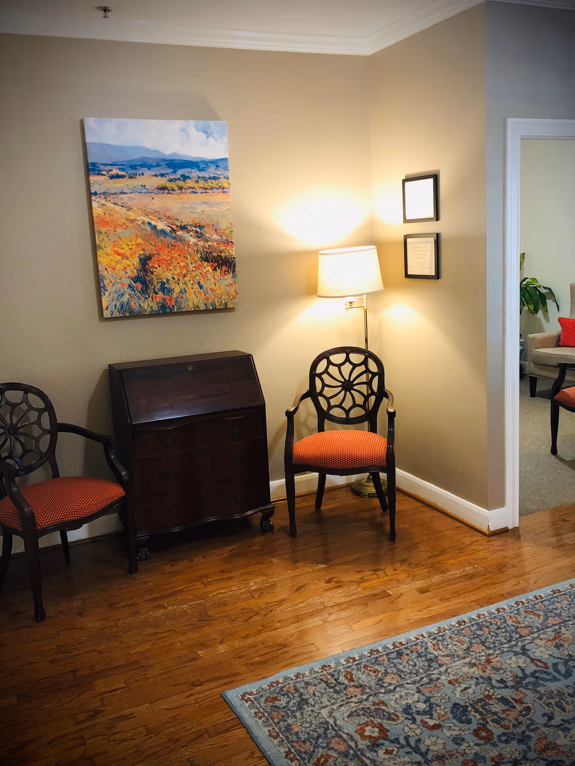 A cozy interior corner of a room with two wooden chairs featuring red cushioned seats, a dark wooden cabinet, a floor lamp emitting warm light, and a colorful landscape painting on the beige wall. A patterned rug covers part of the wooden floor, and an adjoining room with additional seating is visible through an open doorway.