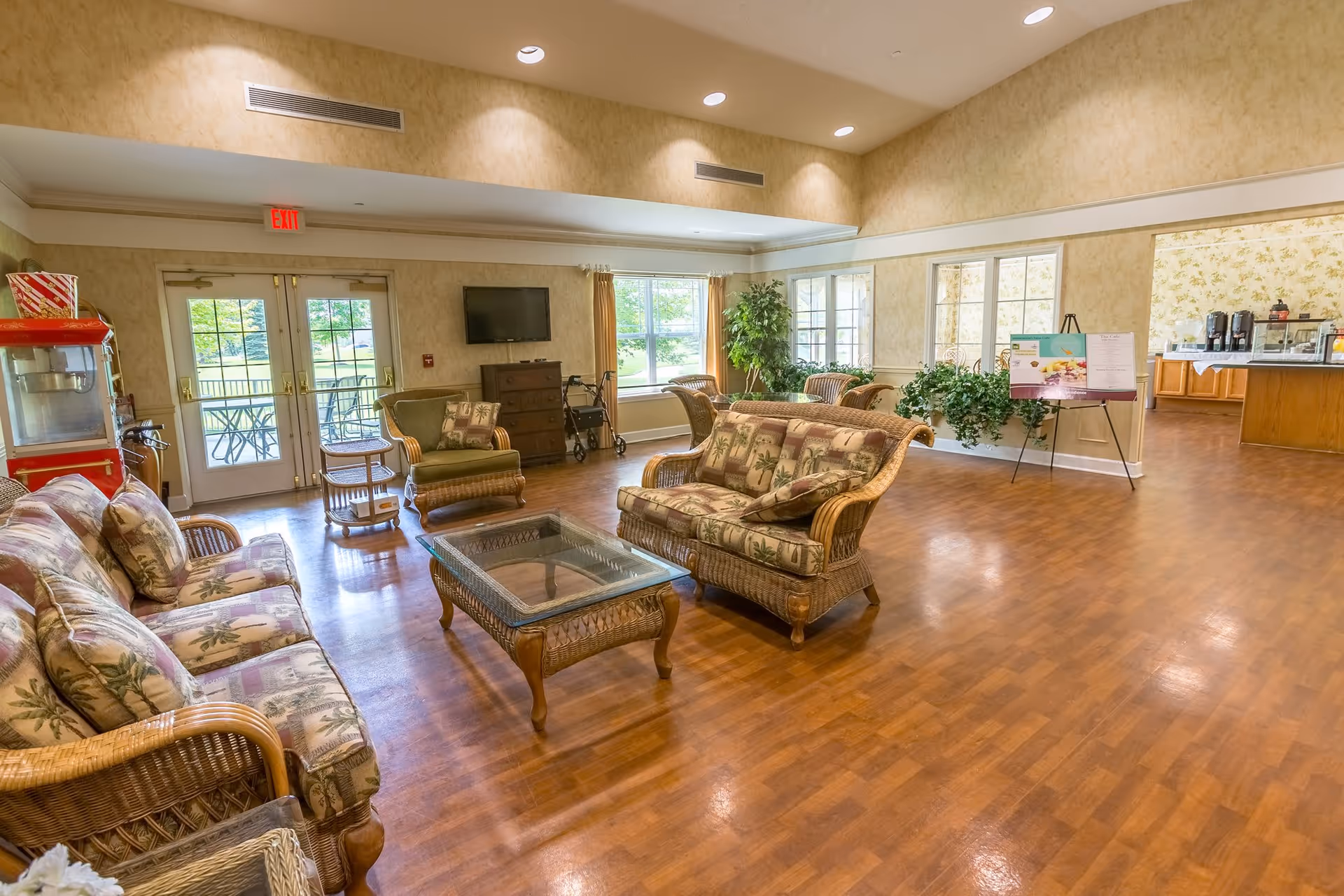 A spacious living room area with wicker furniture including sofas and armchairs with floral cushions, a glass-top coffee table, and a small round side table. There is a popcorn machine on the left near double glass doors leading outside. A flat-screen TV is mounted on the wall above a small cabinet, and a walker is positioned nearby. Large windows let in natural light, and there are several green plants around the room. In the background, there is an open doorway leading to a kitchen or dining area with coffee machines and a counter.