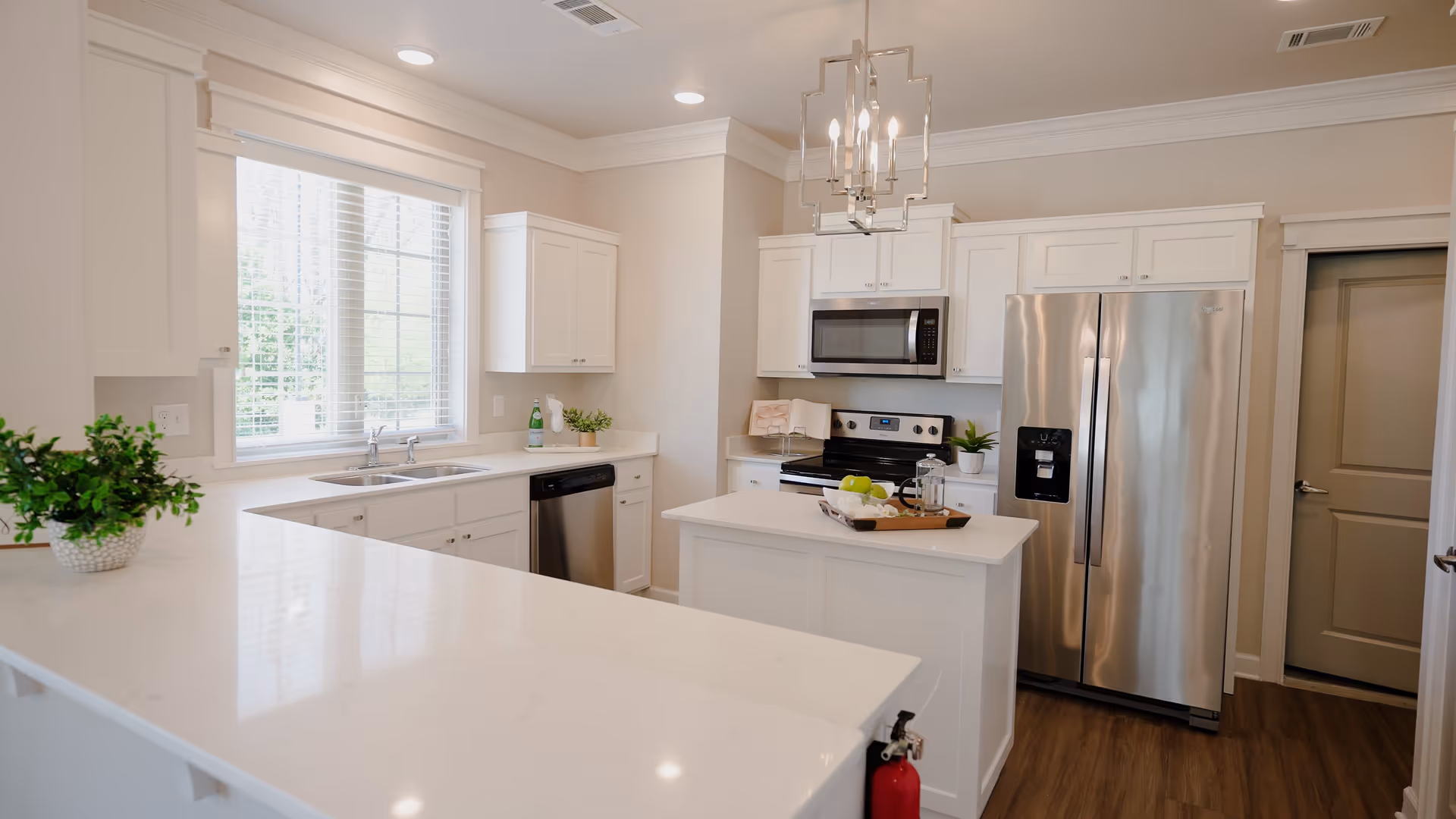 Bright and modern kitchen with white cabinetry, stainless steel refrigerator, microwave, and dishwasher. A small island with a tray holding green apples and a French press is in the center. There is a large window with blinds above the sink, and a contemporary chandelier hangs from the ceiling. The floor is wood, and a fire extinguisher is mounted on the side of the counter.