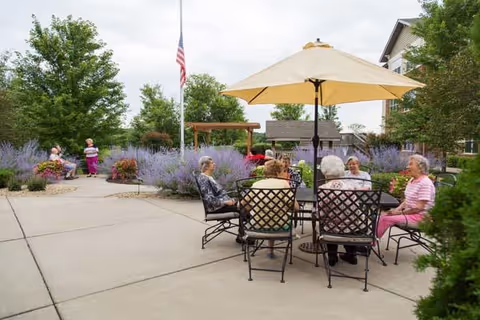 A group of elderly people sitting around a table with a large beige umbrella in an outdoor patio area surrounded by flowering plants and greenery. Two other elderly individuals are walking along a pathway nearby. The setting appears to be a garden or courtyard of a senior living facility with residential buildings in the background and an American flag on a flagpole.