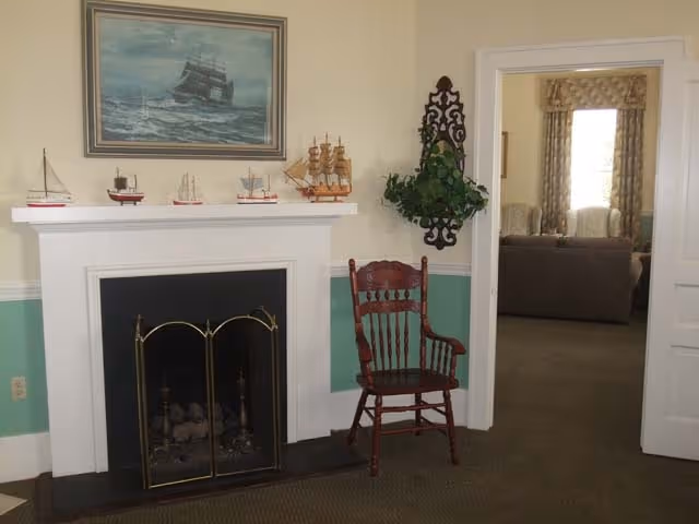 A cozy interior room featuring a white fireplace with a black firebox and brass screen. On the mantel are several small model ships and a framed painting of a sailing ship above it. To the right of the fireplace is a wooden chair with armrests and a decorative wall-mounted plant holder with green foliage. An open doorway leads to another room with two armchairs and a sofa, with windows covered by patterned curtains.
