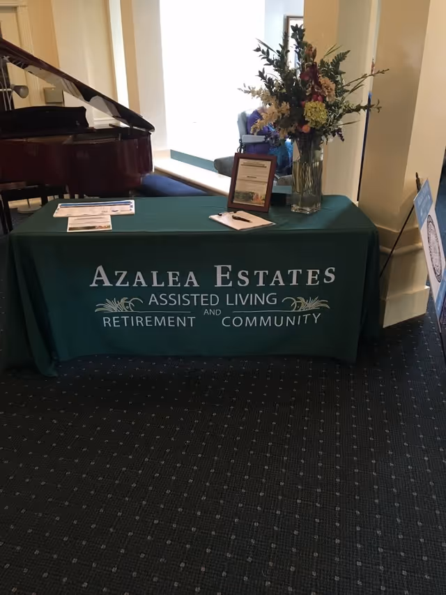 A lobby table draped with a green 'Azalea Estates Assisted Living and Retirement Community' cloth, holding a vase of flowers, brochures and a clipboard with a grand piano in the background.