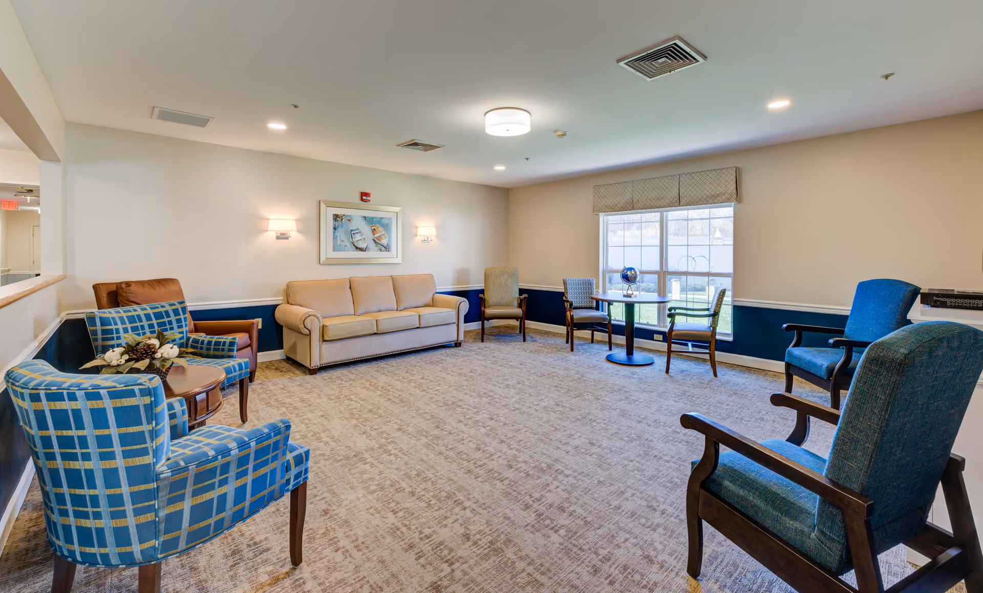 A spacious and well-lit common area with multiple upholstered chairs and a beige sofa arranged around the room. There is a small round table with a globe near a large window letting in natural light. The walls are painted in a light color with a dark blue wainscoting, and a framed picture hangs above the sofa. The carpet is light-colored and the ceiling has recessed lighting and a central light fixture.