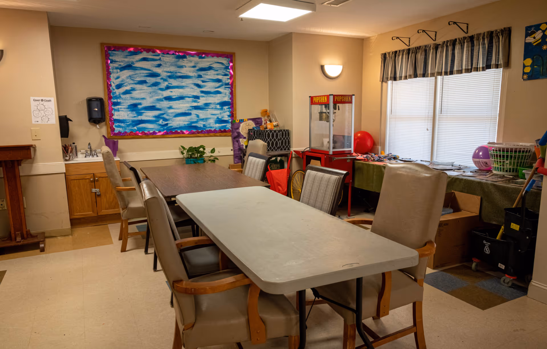 A senior living facility activity room with two tables surrounded by chairs. There is a popcorn machine on a red cart near the window with closed blinds. The walls are beige with a bulletin board featuring a blue and white abstract design. Various items, including a plant, cleaning supplies, and craft materials, are placed around the room.