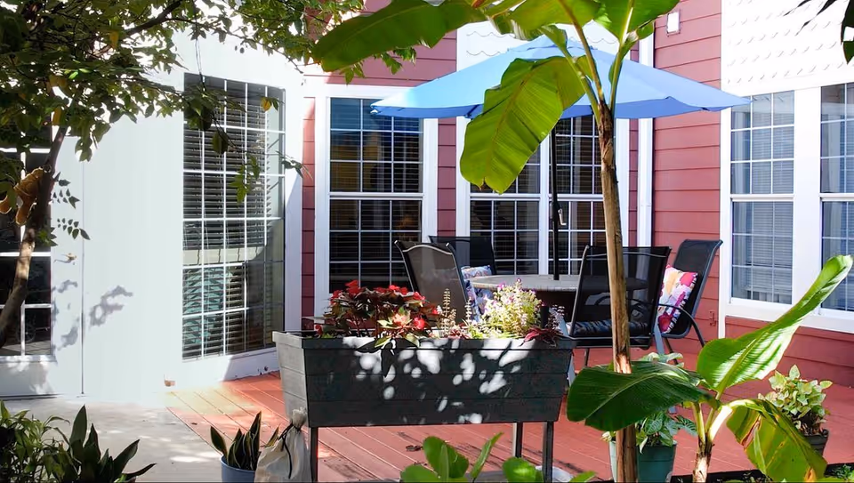 Outdoor patio area with a wooden deck, potted plants, and a table with chairs under a blue umbrella. The background shows red siding walls with multiple windows and a glass door.