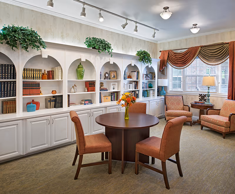 A cozy living room area with a round wooden table surrounded by four upholstered chairs in a warm orange fabric. Behind the table is a large white built-in bookshelf with decorative items, books, and green plants on top. To the right, there are two upholstered armchairs with a small round wooden side table holding a lamp and decorative items. A large window with elegant drapes allows natural light into the room.