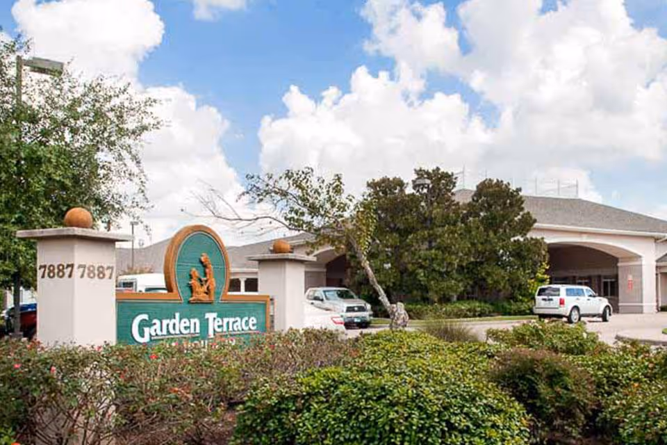 Entrance sign reading 'Garden Terrace' in front of the facility with landscaped shrubs, driveway, cars, and the building under a partly cloudy sky.