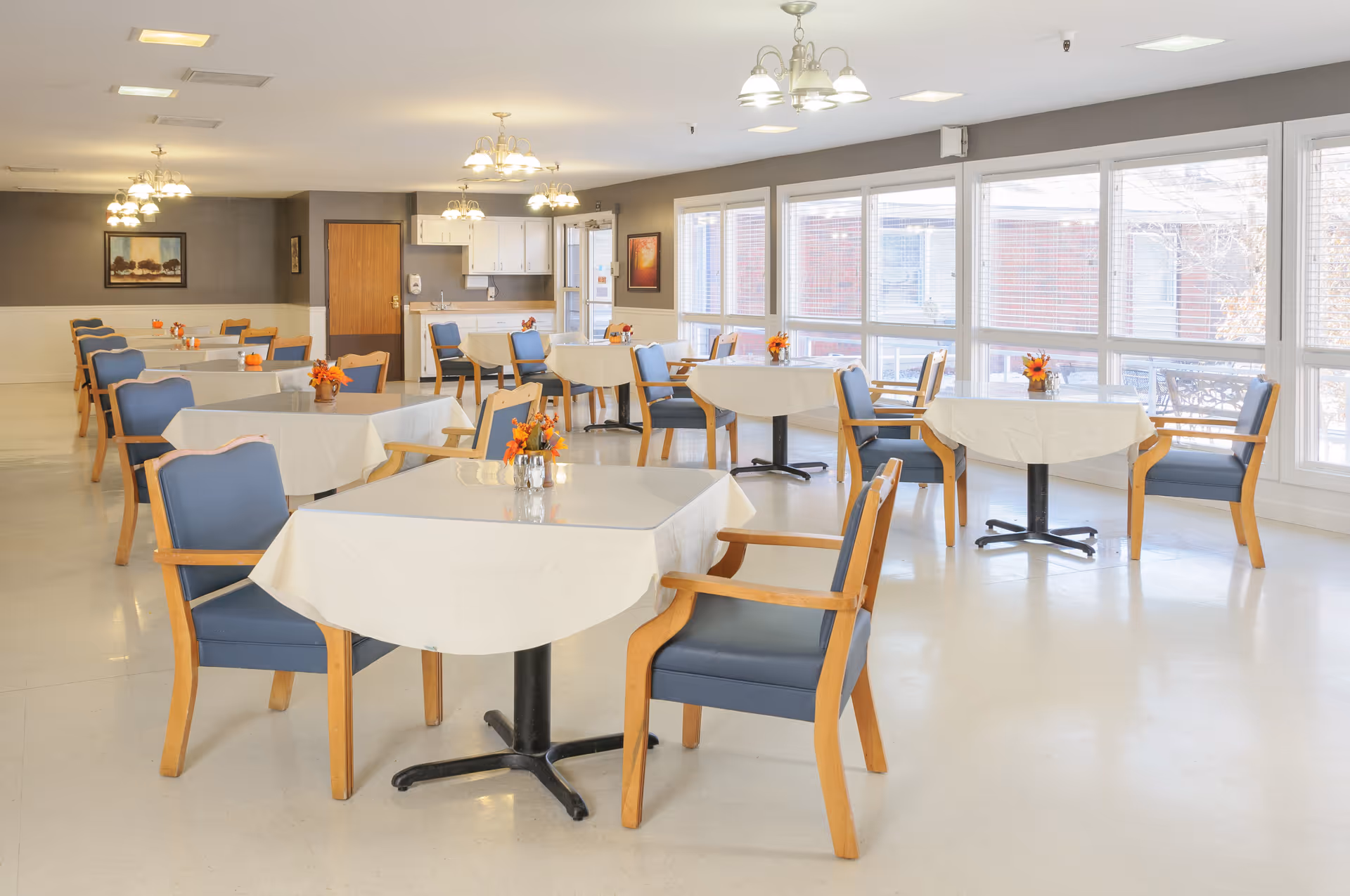 A bright dining room with multiple tables covered with white tablecloths, each surrounded by wooden chairs with blue cushions. Small floral centerpieces are placed on each table. Large windows on one side allow natural light to fill the room. The room has light-colored walls and ceiling with several ceiling light fixtures.