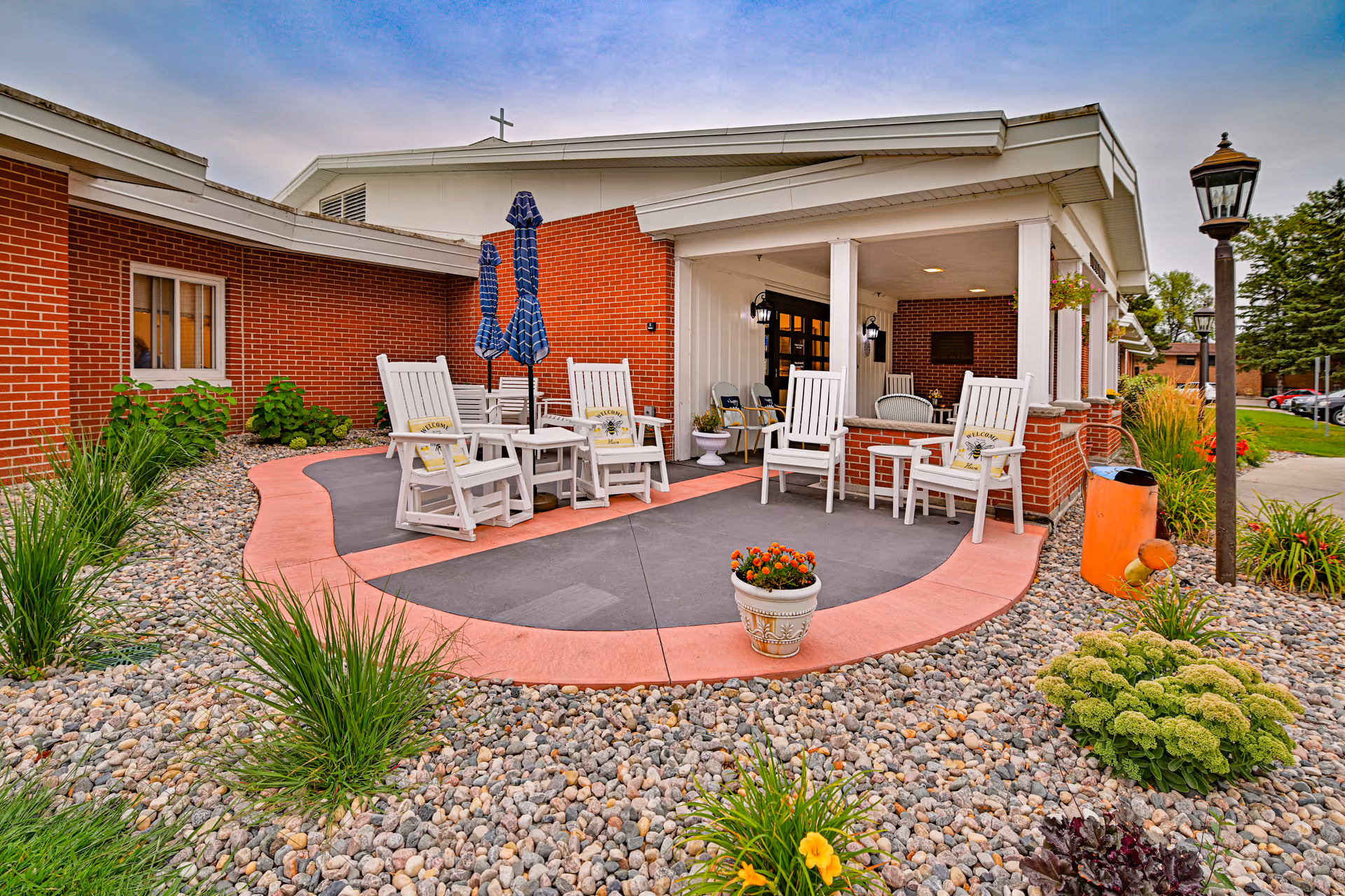 Outdoor patio area at Valley Senior Living on Cherry - Tufte Manor featuring white wooden rocking chairs with 'Welcome' cushions, small tables, closed blue umbrellas, surrounded by decorative rocks and plants, adjacent to a brick building with a covered porch and a lamppost nearby.